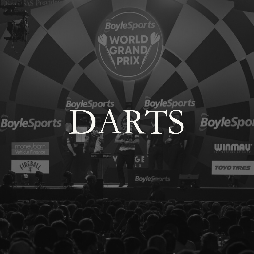 A stage setup at a darts event with the BoyleSports World Grand Prix logo overhead, and the word "DARTS" displayed prominently in the center. Several people are on stage, and a large audience is watching.