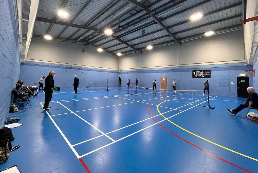 People playing pickleball in an indoor gymnasium with blue walls and a high ceiling.