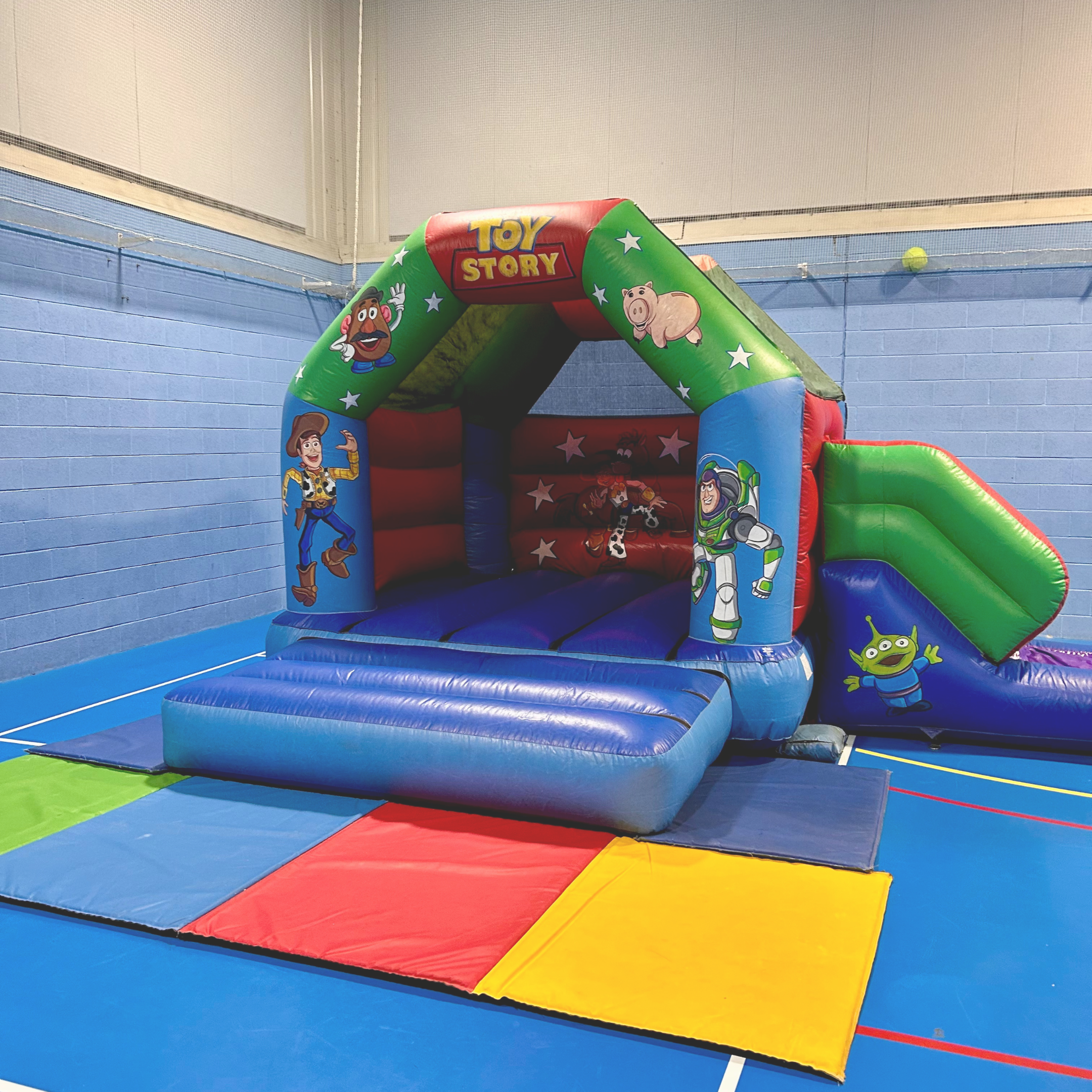 Colorful inflatable bouncy castle featuring Toy Story characters, set up indoors on blue mats in a gym.