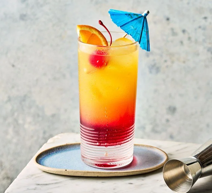 Colorful layered cocktail in a tall glass with garnish of orange slice, maraschino cherry, and blue paper umbrella