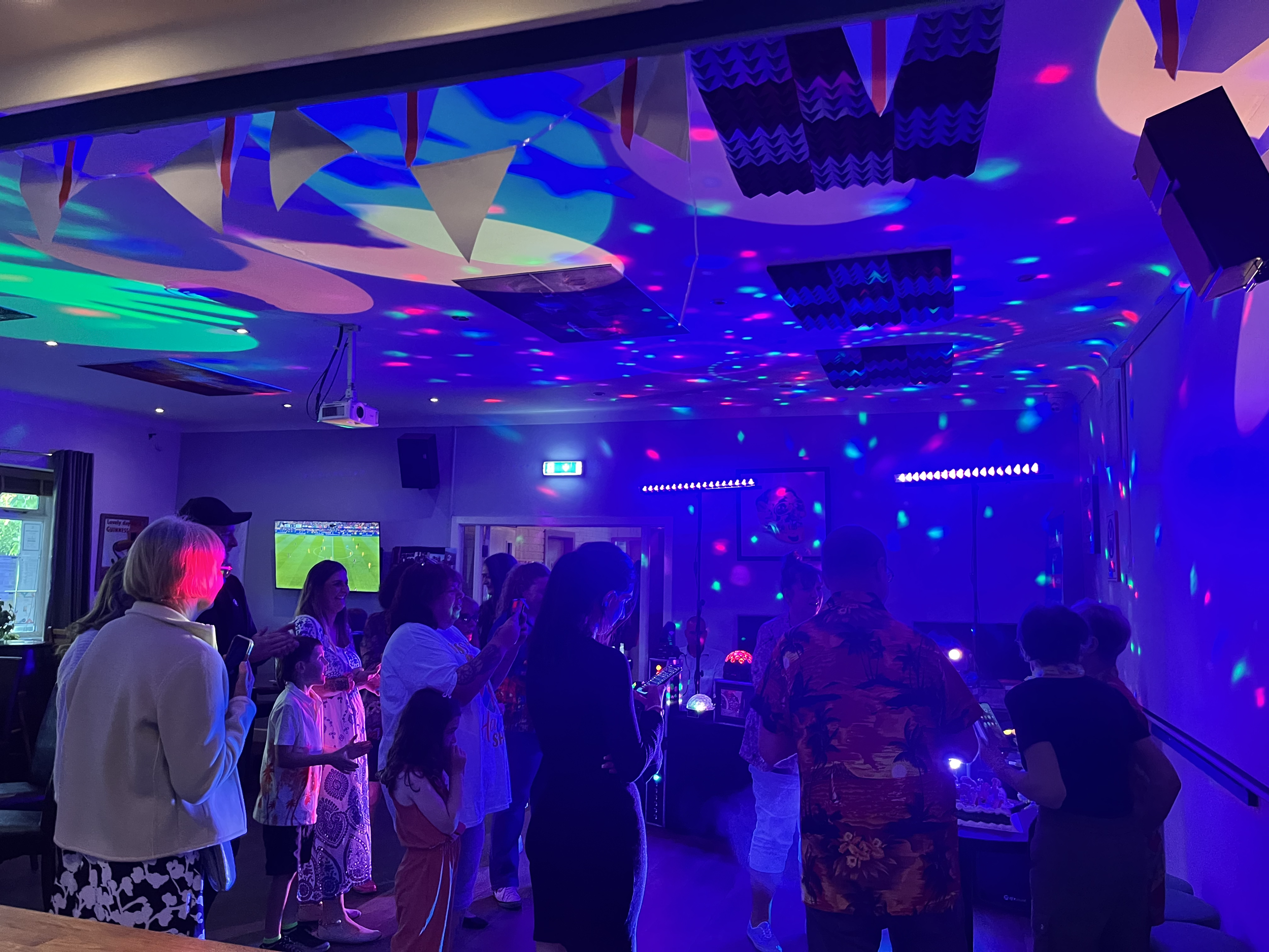 People gathered in a dark room with colorful disco lights overhead, standing around a table with various lights or decorations, while some are looking at their phones and others are chatting.
