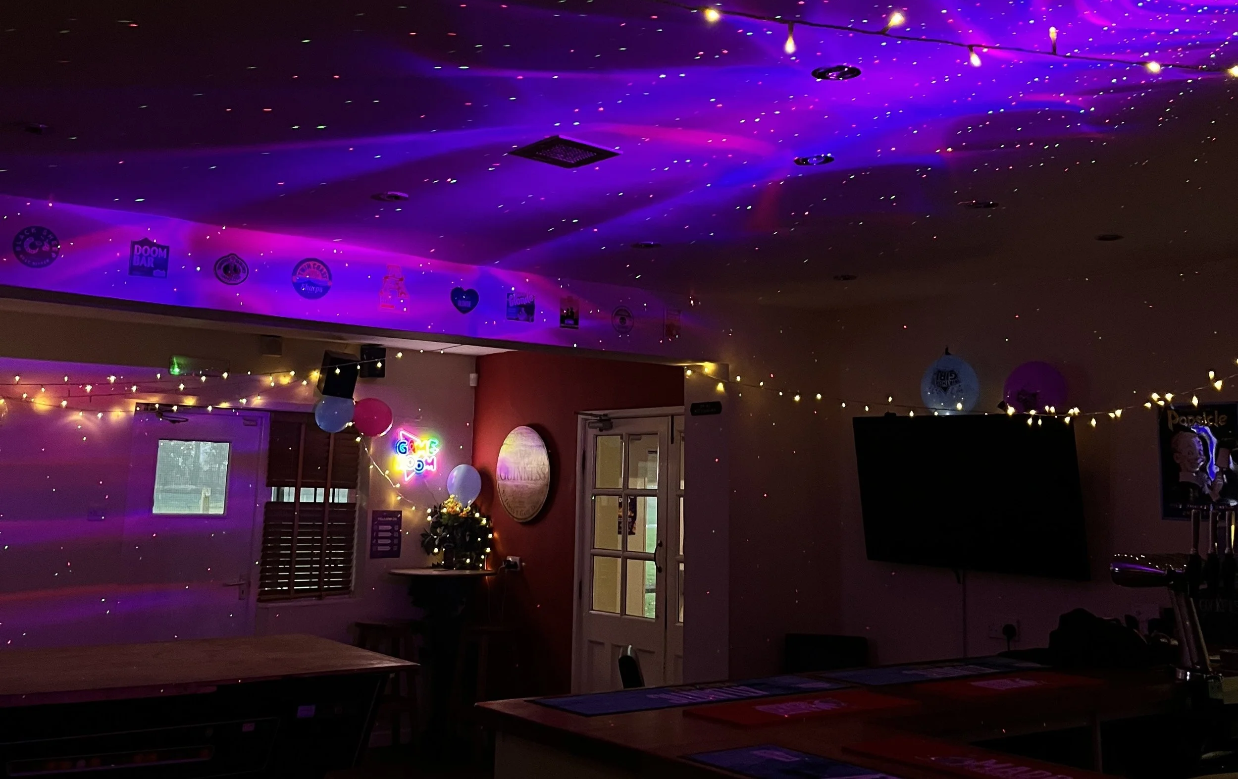 Party room decorated with colorful string lights, balloons, and wall banners, with a mounted TV and a neon sign that says 'Game On'.