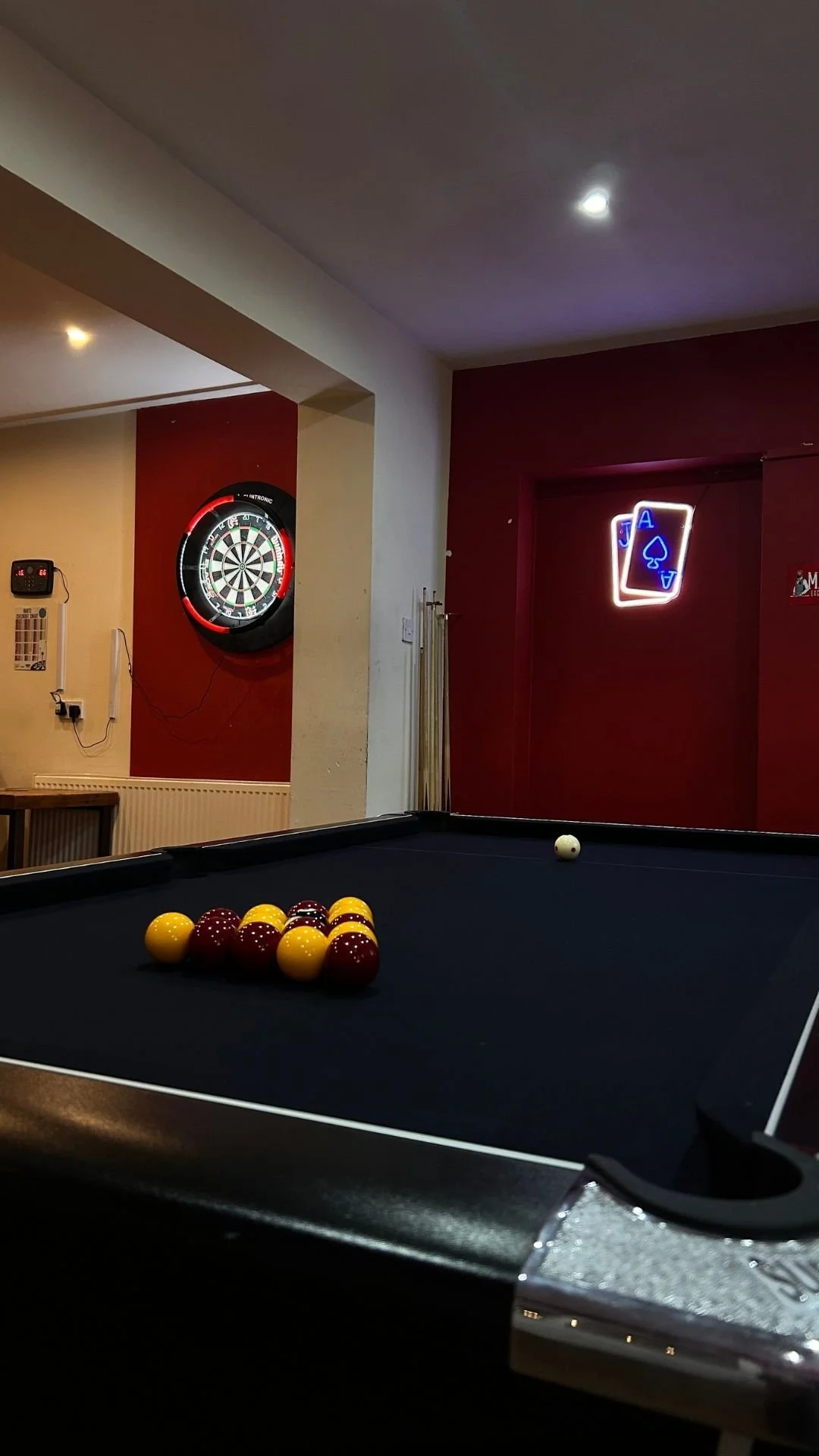 A pool table with yellow and maroon billiard balls arranged in a triangle and a white cue ball on a dark playing surface in a recreational room. In the background, there is a dartboard and neon signs, including playing cards and an ace of spades.