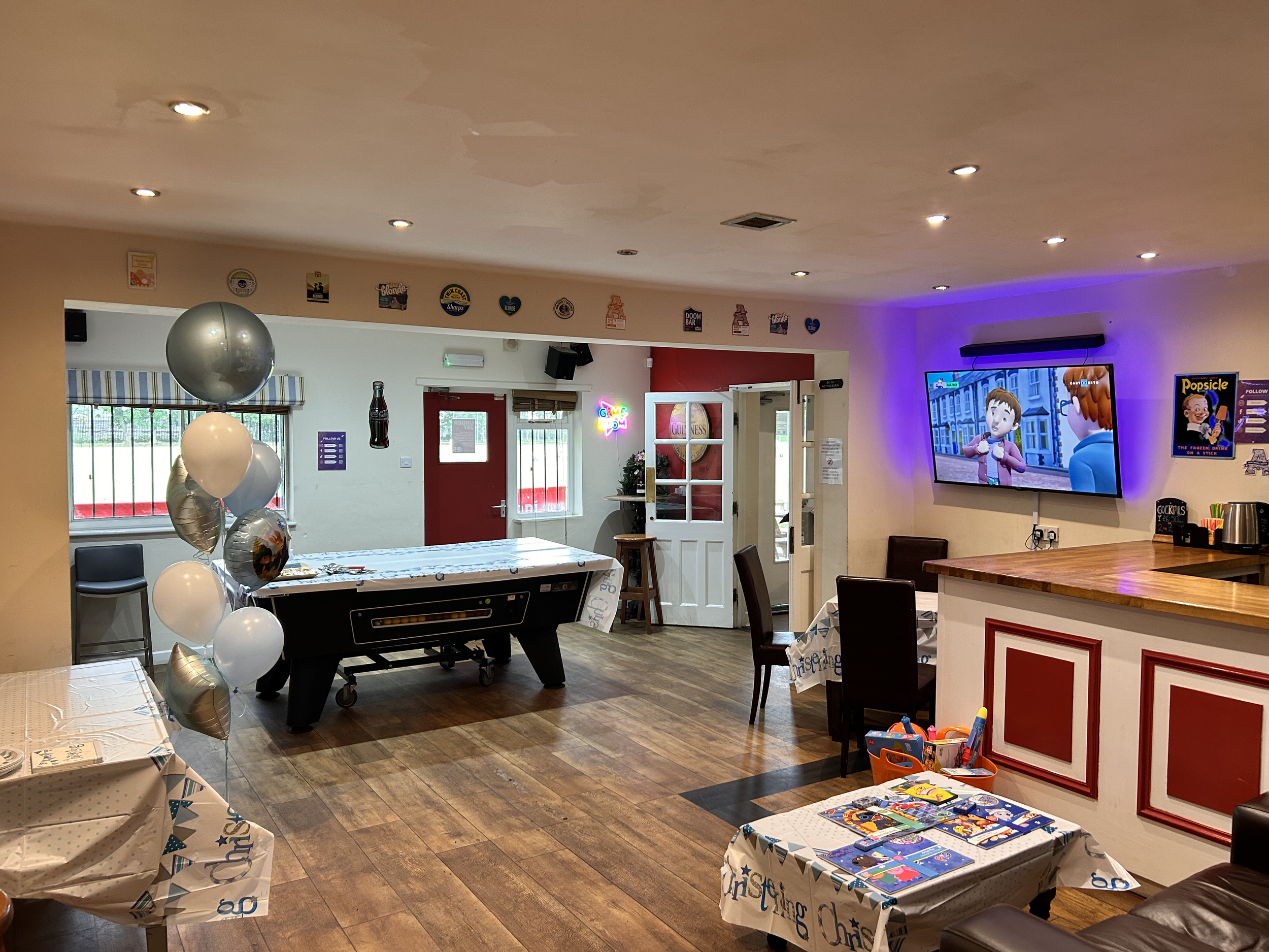 A decorated party room with balloons, a table with party supplies, a air hockey table, a television displaying animated characters, and party-themed decorations on the walls.
