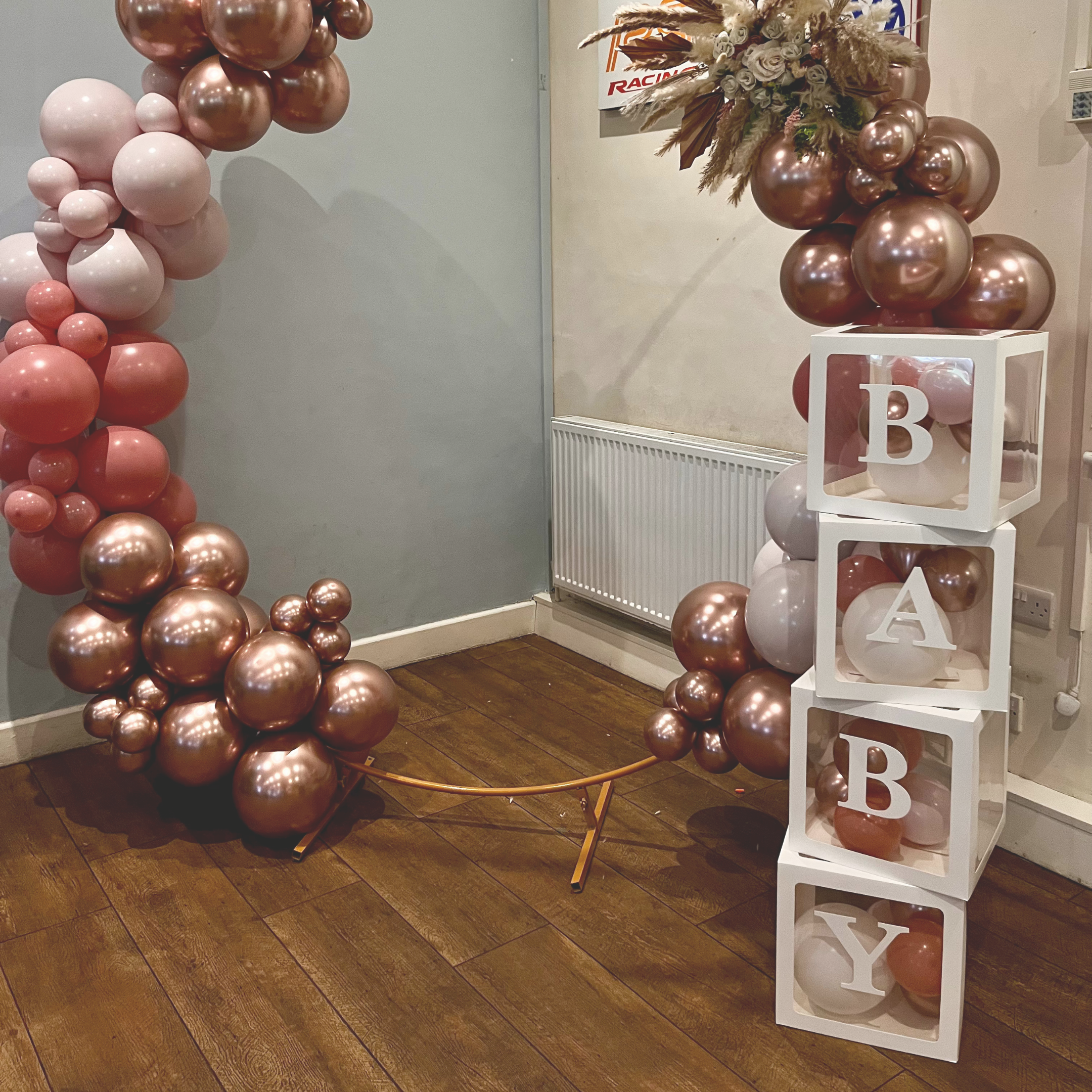 Decorative balloon arrangement with pink, white, and rose gold balloons, including a white cube display with the letters B, A, B, and Y, forming the word 'BABY.'