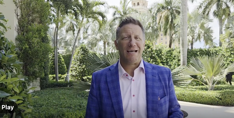 A man in a blue plaid blazer and white shirt standing outdoors among palm trees and lush greenery, making a facial expression as if surprised or confused.