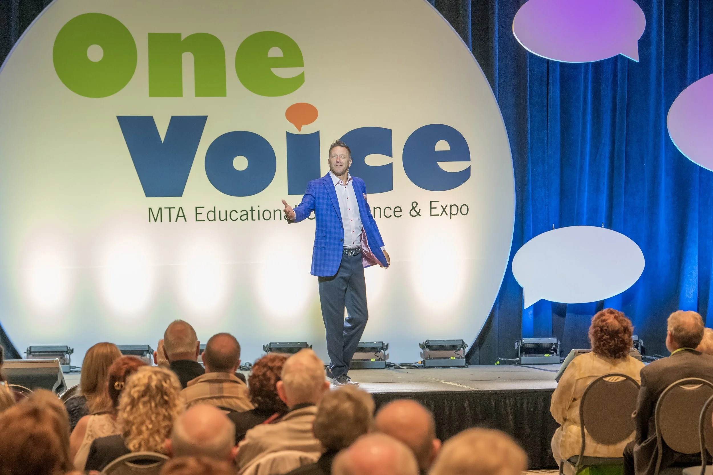 A man in a blue checkered blazer and dark pants stands on stage in front of a large screen that reads "One Voice" and "MTA Education Conference & Expo." He is addressing an audience, with several people in the front rows listening attentively.