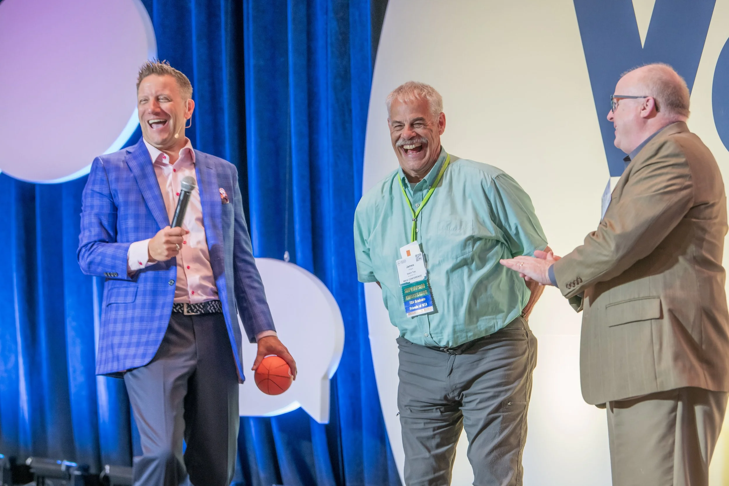 Three men smiling and laughing on stage at a conference or event, with one holding a microphone and a basketball, and wearing name tags.
