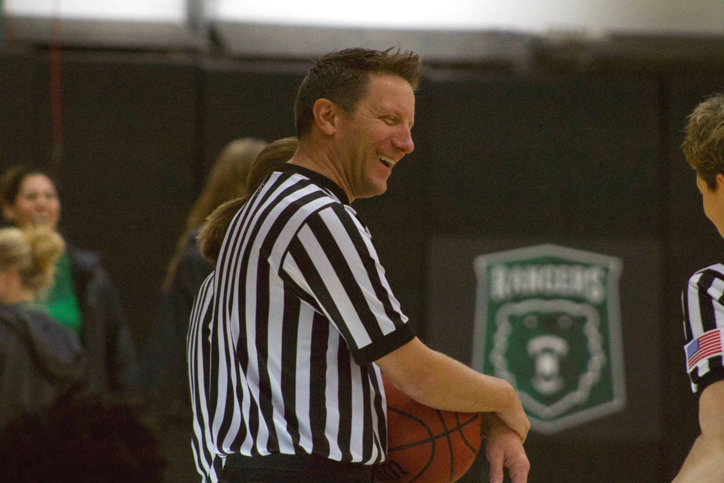 A man wearing a black-and-white striped referee shirt holding a basketball, smiling while talking to a woman, with other people and a green sports emblem in the background.