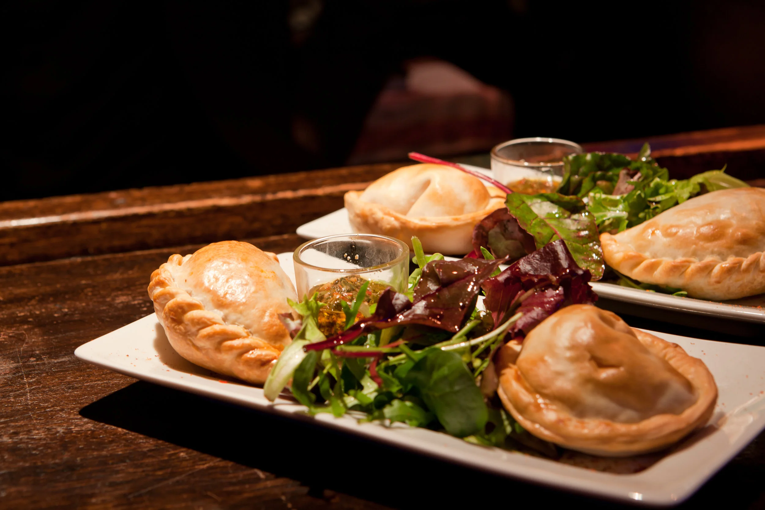 Plat d'empanadas, spécialité Argentine, accompagnées d'une salade verte avec vinaigrette et deux petits pots de sauce chimichuri ou dips, présenté sur une assiette blanche sur une table en bois.