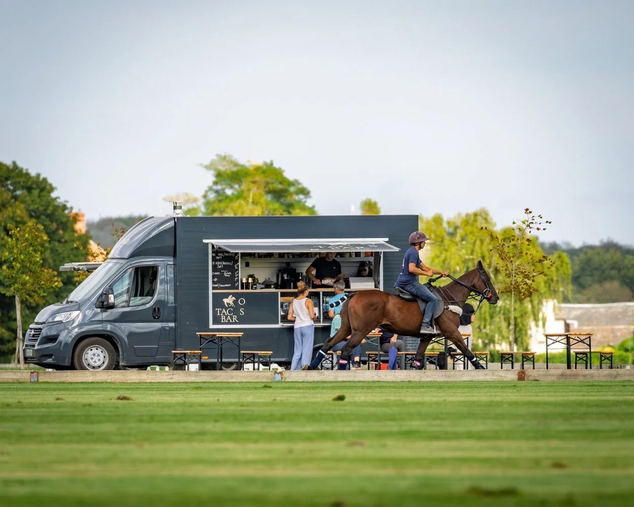 Le Tacos Bar, food-trucks du club , Un camion de nourriture converti en bar avec des clients commandant au bord d'une terrain de polo, un homme à cheval passant devant le camion, et un environnement vert avec des arbres en arrière-plan