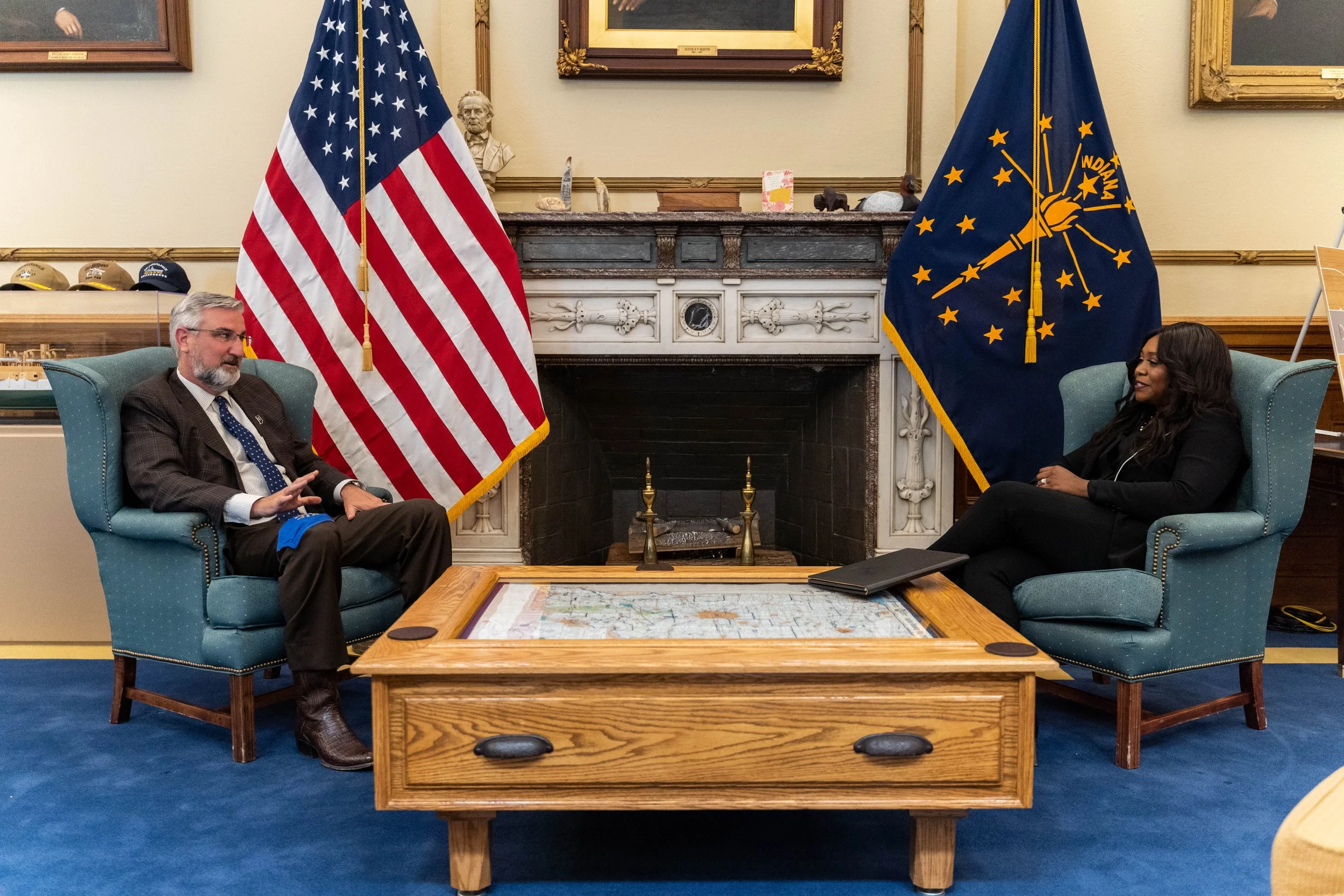 Two people sitting in armchairs facing each other in a room with an ornate fireplace, flags of the United States and Indiana in the background, and a framed portrait on the wall. One man, with gray hair and beard, wearing a suit, and a woman with long dark hair, wearing dark clothing.