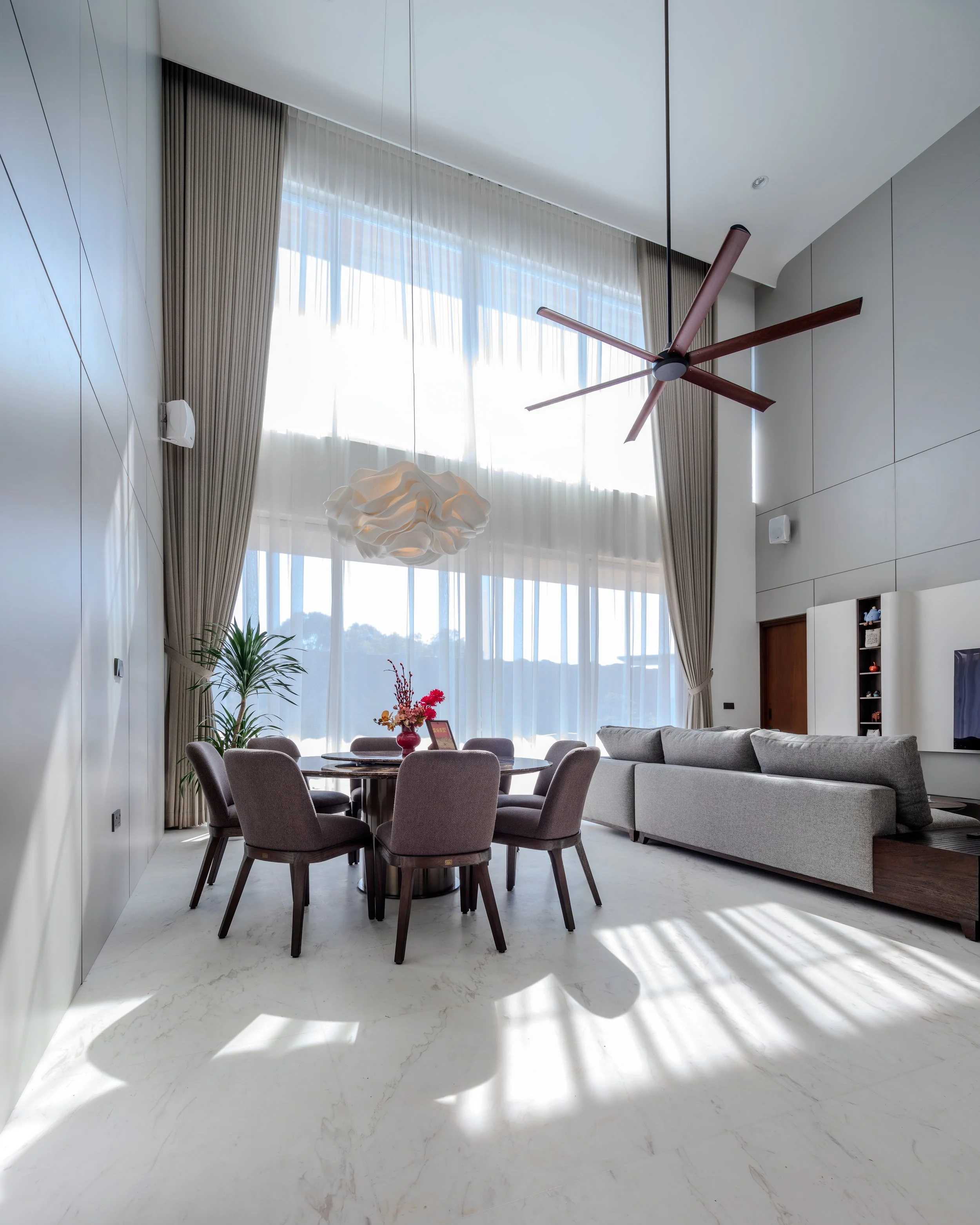 Living room with high ceiling, large windows with sheer curtains, a round dining table with six chairs, a modern chandelier, a ceiling fan, a gray sofa, and minimal decor.