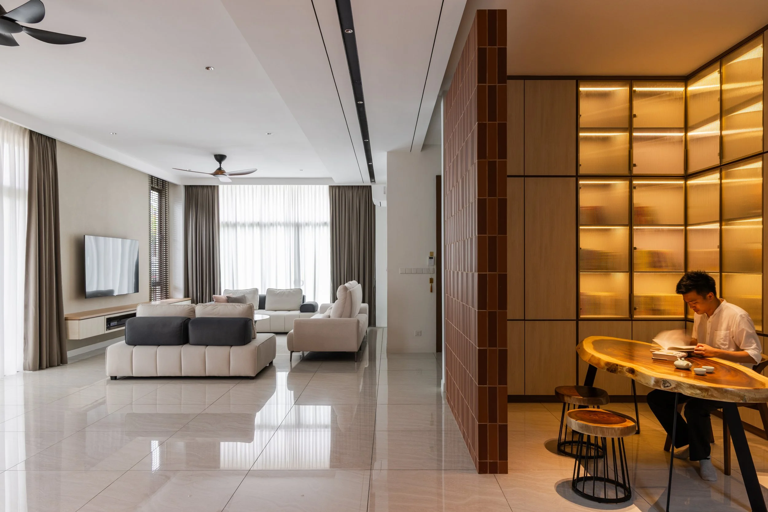Modern living room with beige sofas, a wall-mounted TV, and large windows with curtains. To the right, a man in a white shirt reads a book at a wooden table under warm lighting.