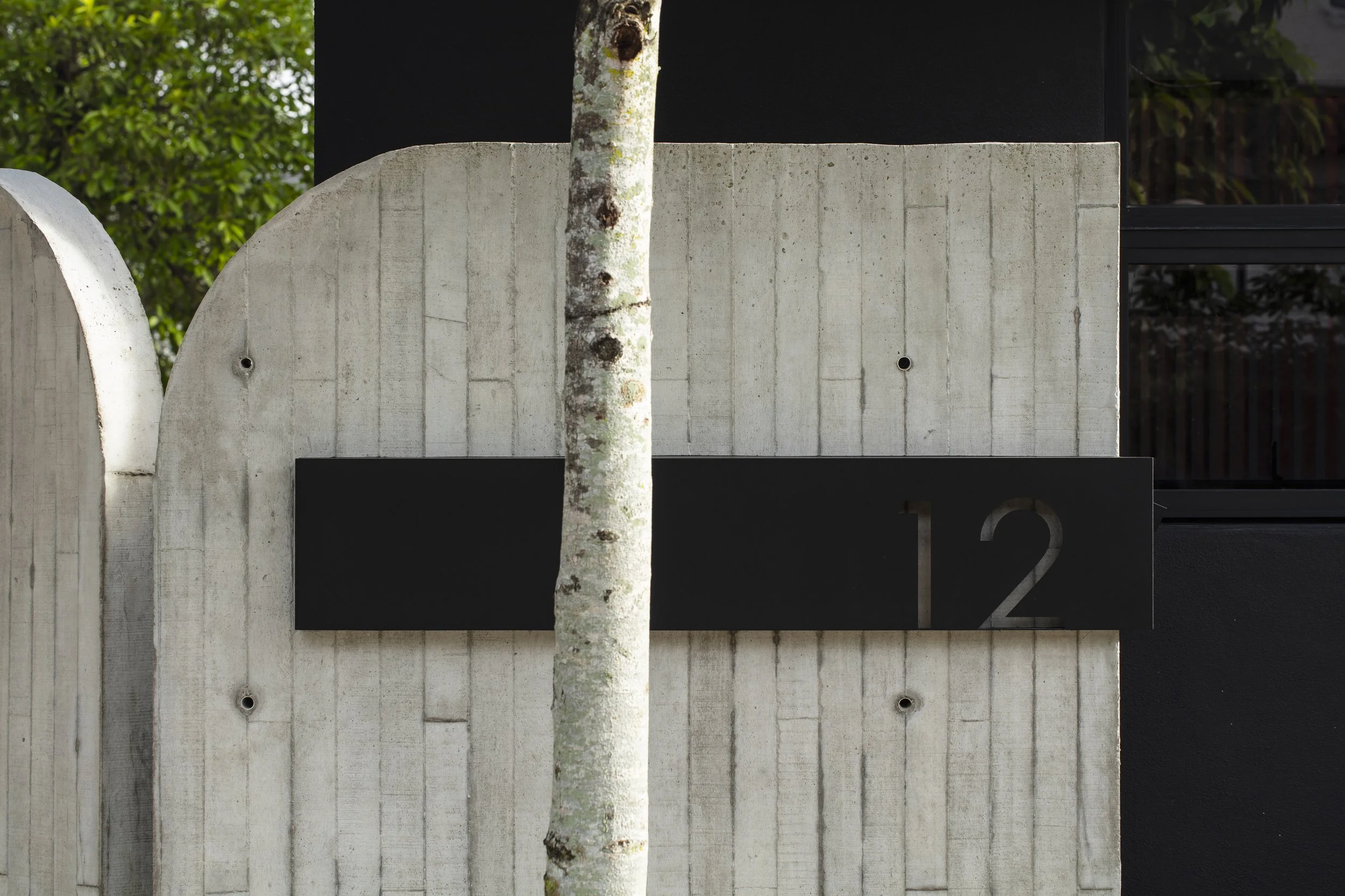 Close-up of a modern house exterior with a concrete wall, black metal numbers '12', a tree trunk in front, and a black window with grill on the side.