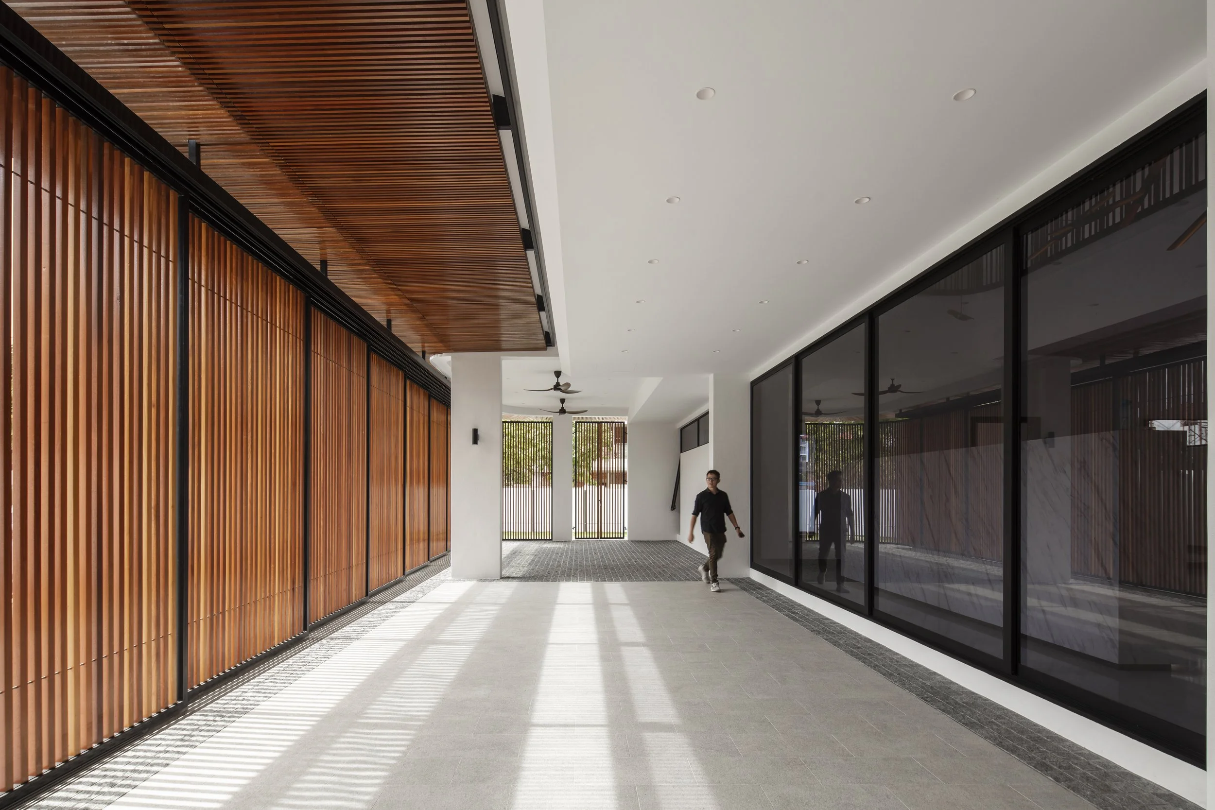 Modern indoor corridor with wooden wall panels on the left, large glass windows on the right, white ceiling with recessed lighting, and a person walking in the distance.