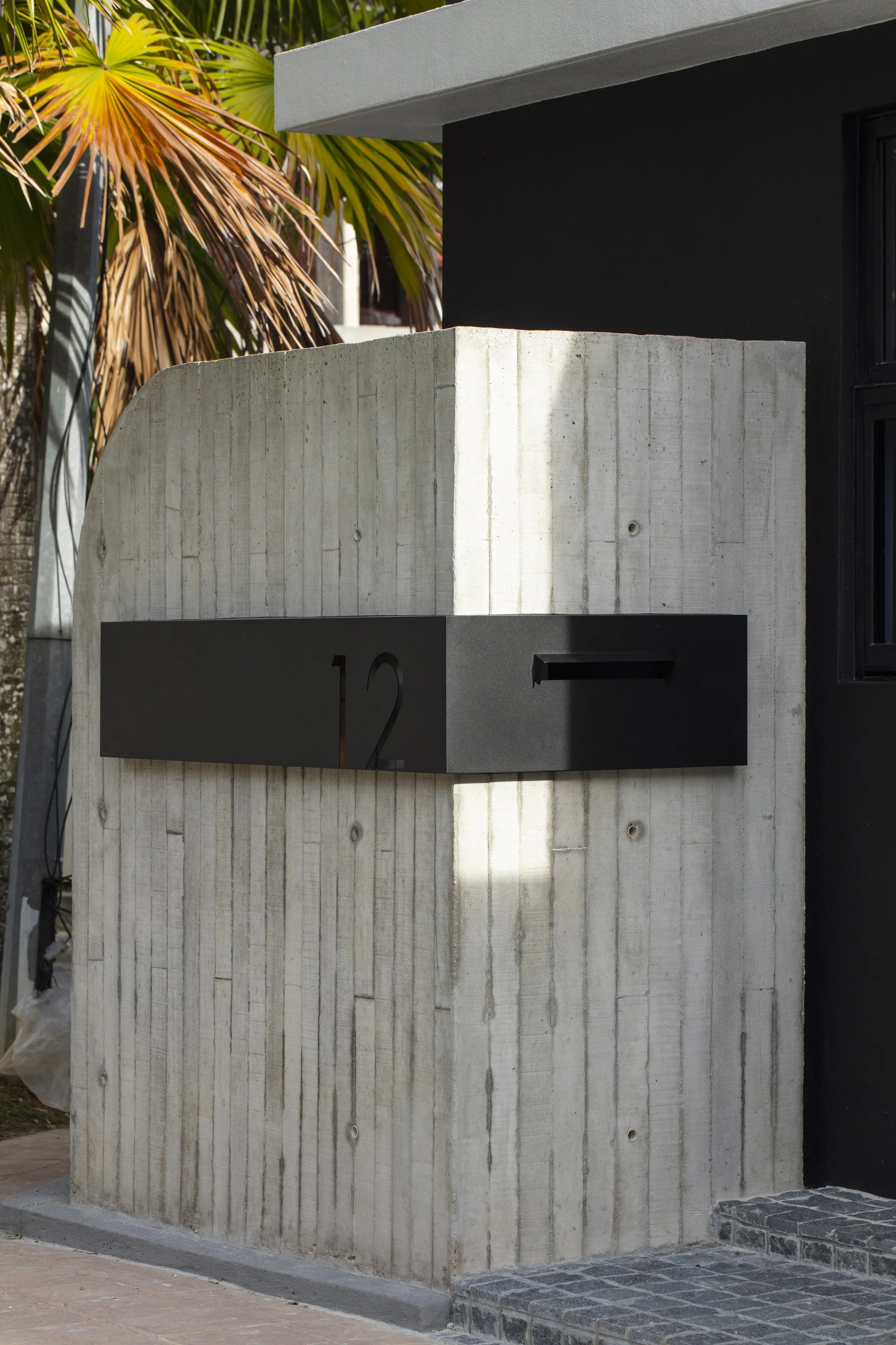 Modern concrete house number wall with black mailbox and house number 12, next to a pathway, with palm trees in the background.