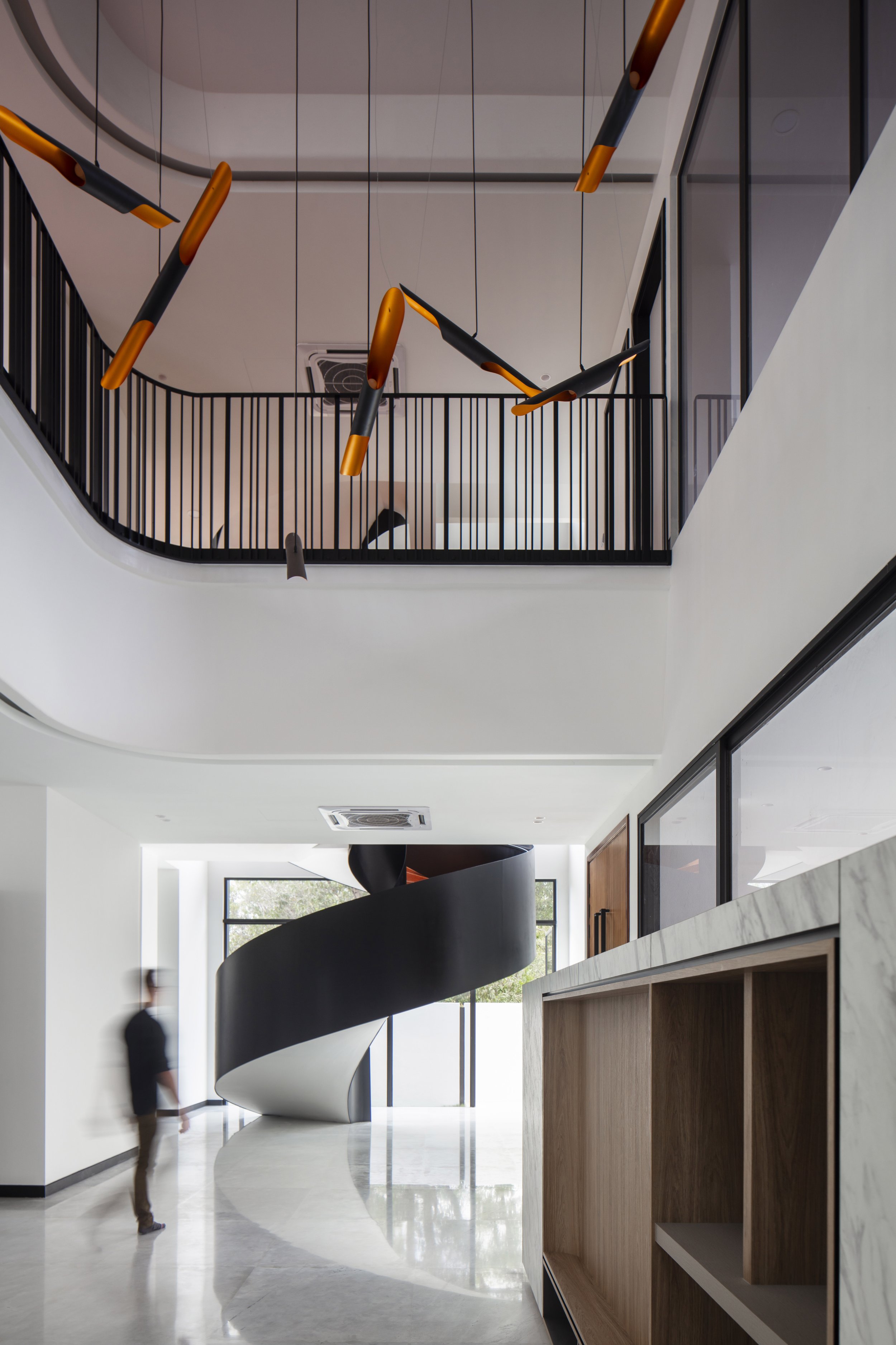 Interior view of a modern building featuring a black spiral staircase, large windows, white walls, and a series of hanging orange and black pendant lights.