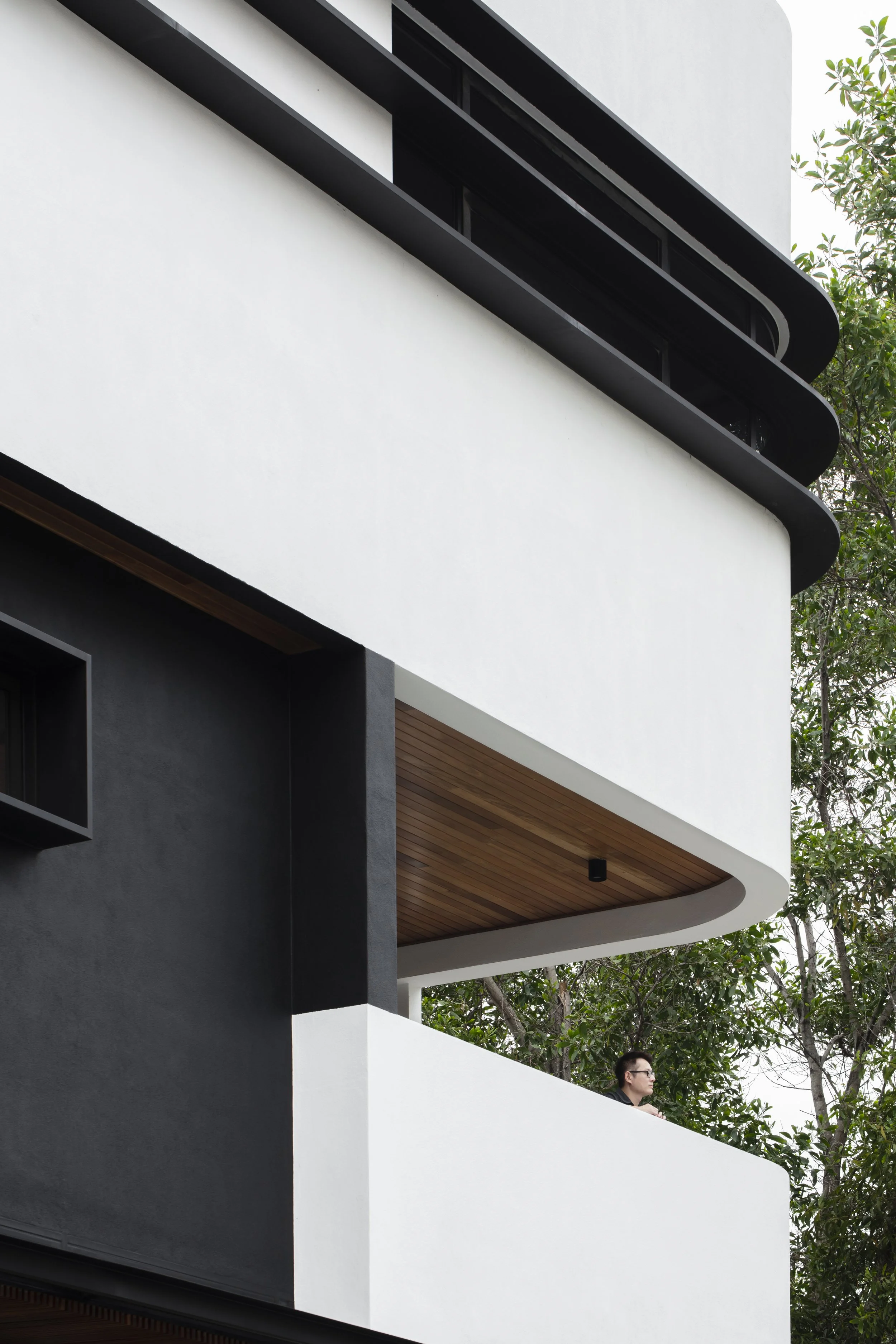 Close-up of a modern building with white and black exterior walls, curved architectural features, and a balcony with a person standing on it, surrounded by green trees.