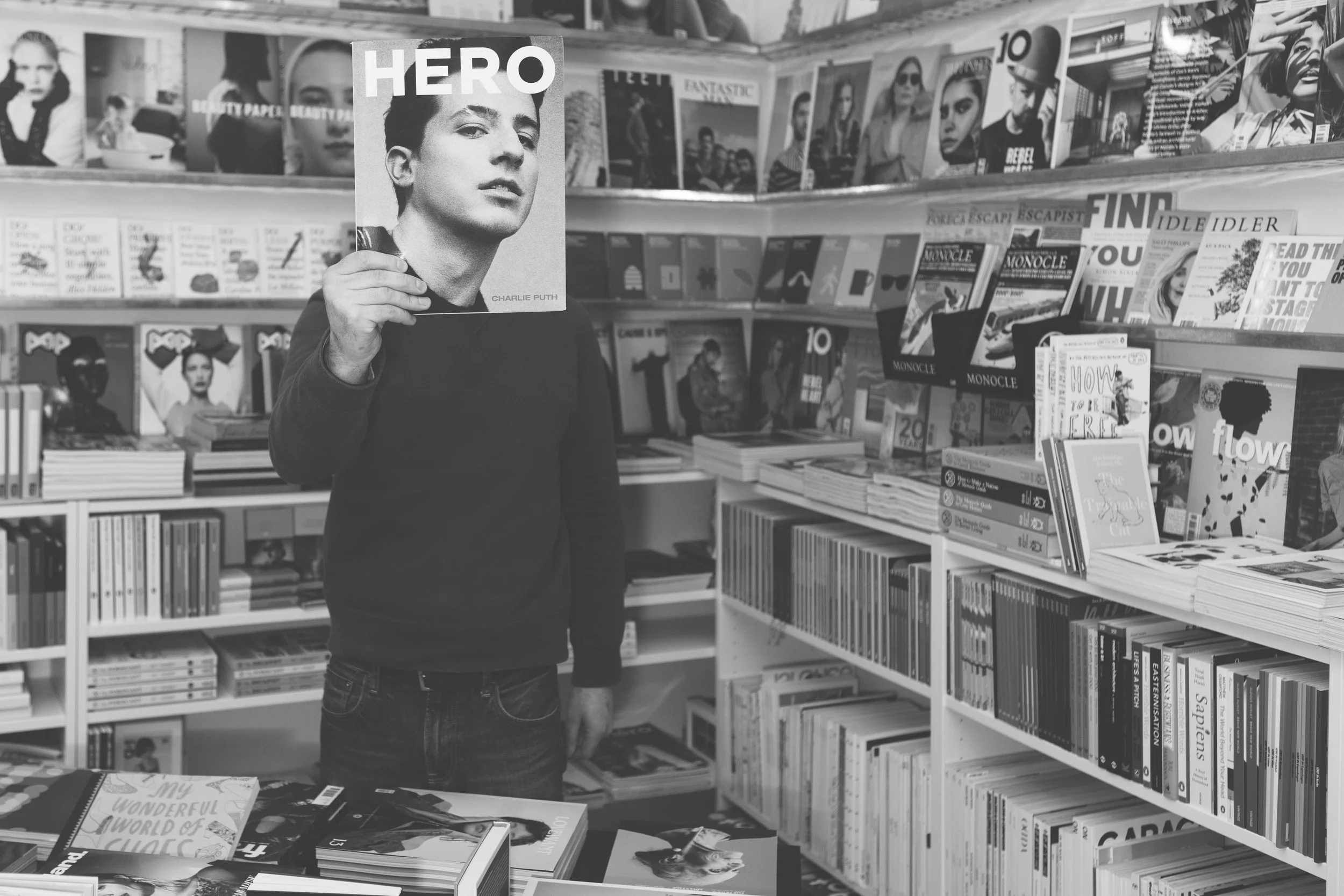 black and white portrait of a man holding a magazine up to his face, with the front cover replacing his face. Surrounded by books either side on white and metal shelves, filled with books and magazines up and down the walls. 