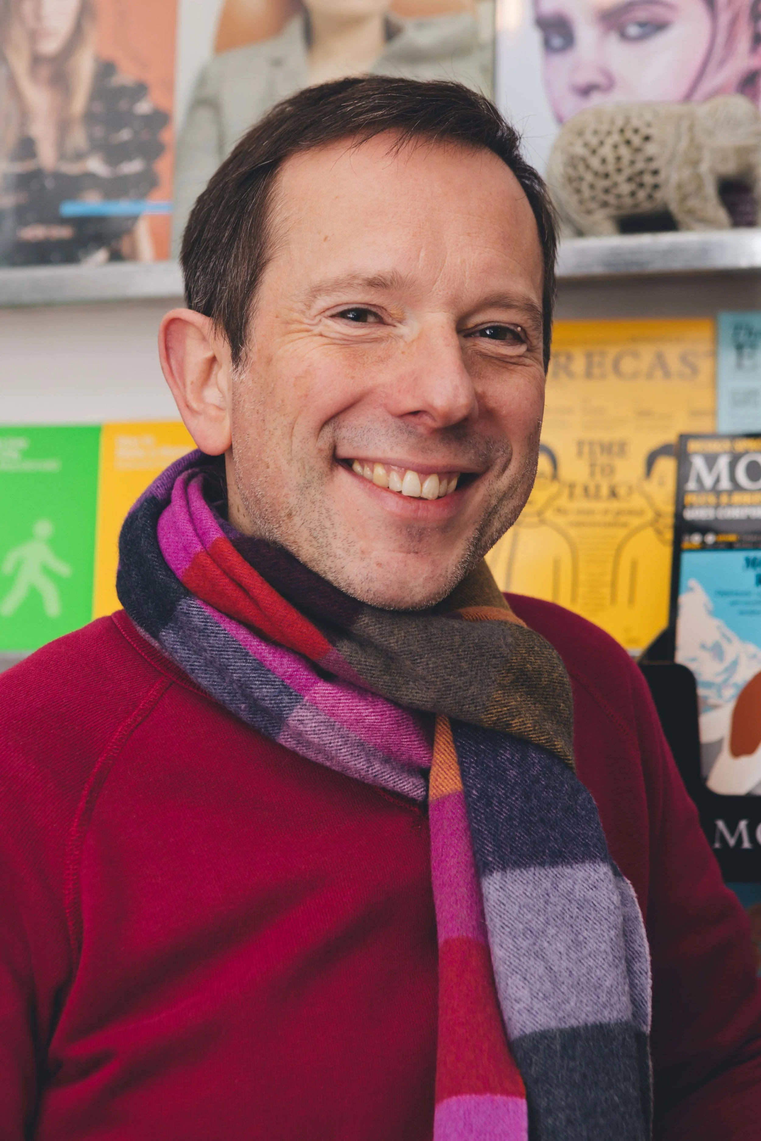 colour portrait image of a man in a red jumper with a striped scarf on smiling to camera, with shelves of books behind him