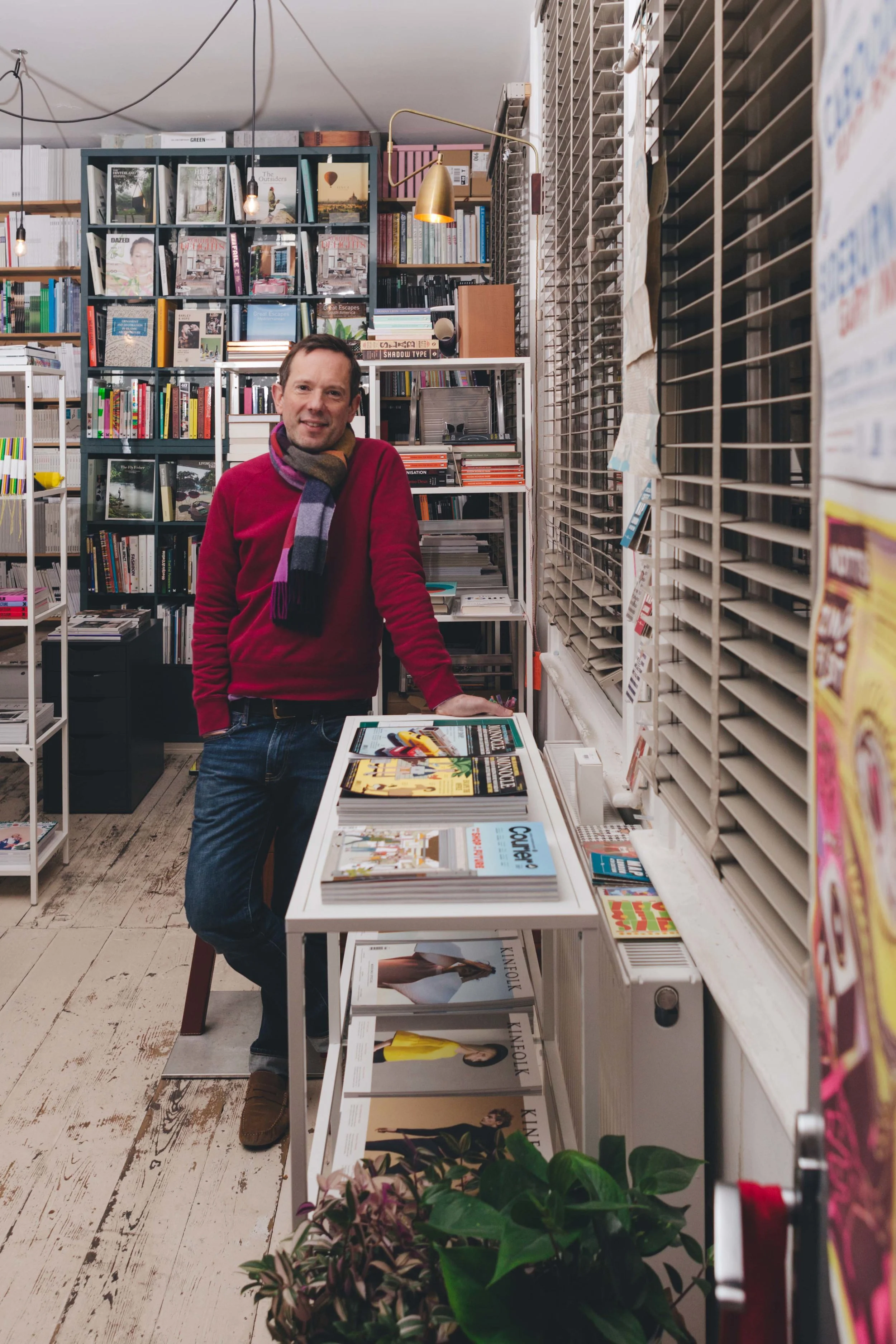 portrait of a man in a book shop with plants in the foreground to the right, blinds also to the right and books in shelves on the walls