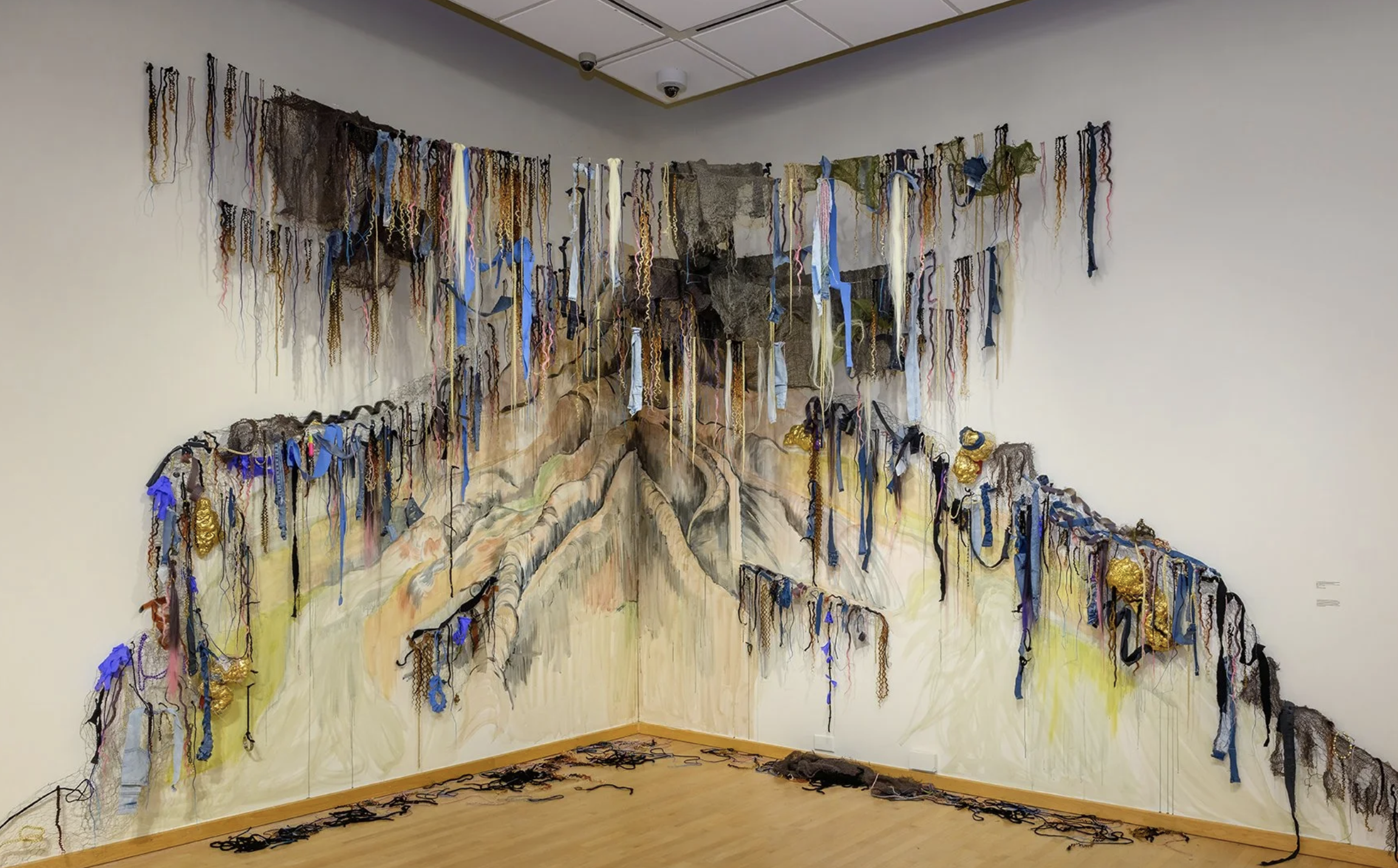 An installation of ribbons, hair, and string crossing the corner of a gallery wall. The roots of a tree are painted on the wall behind it.