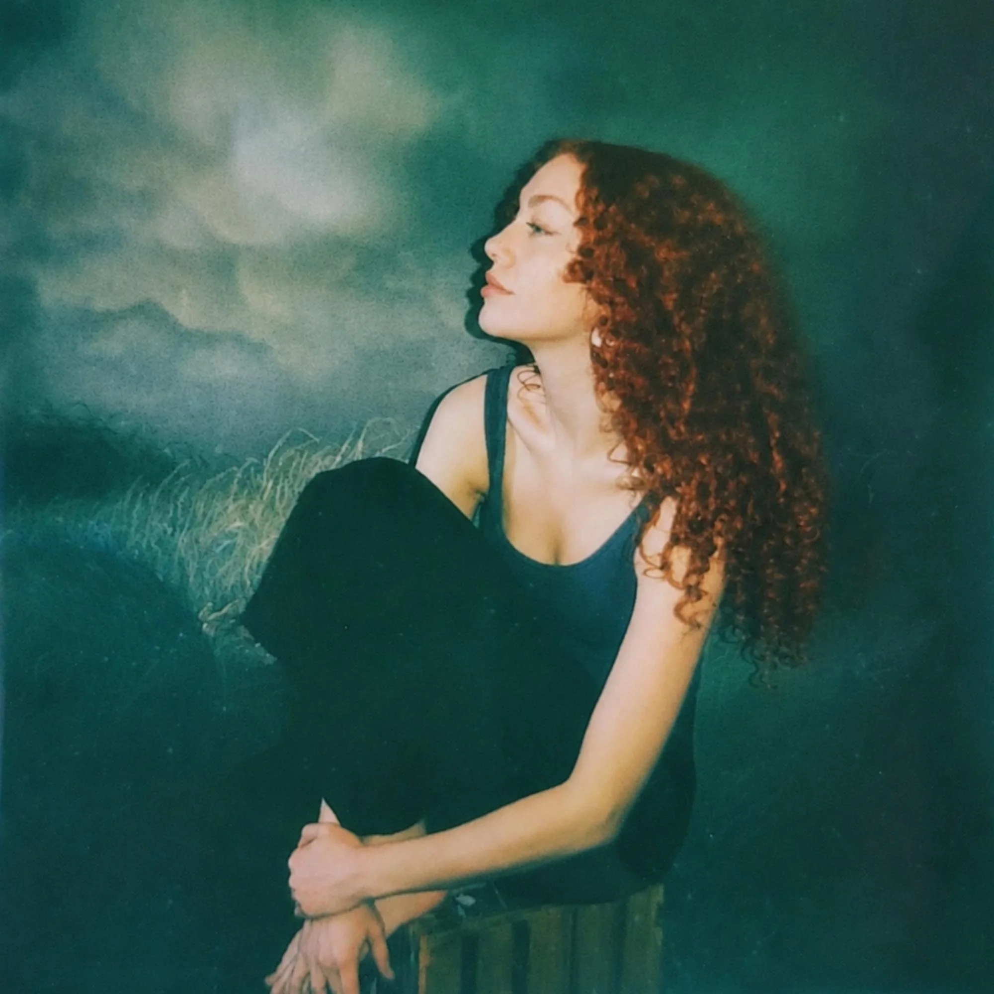 Artist Elizabeth Bergeland, who has red curly hair and wearing a green tank top is looking to the left while knealing in front of a painting of a stormy sky.