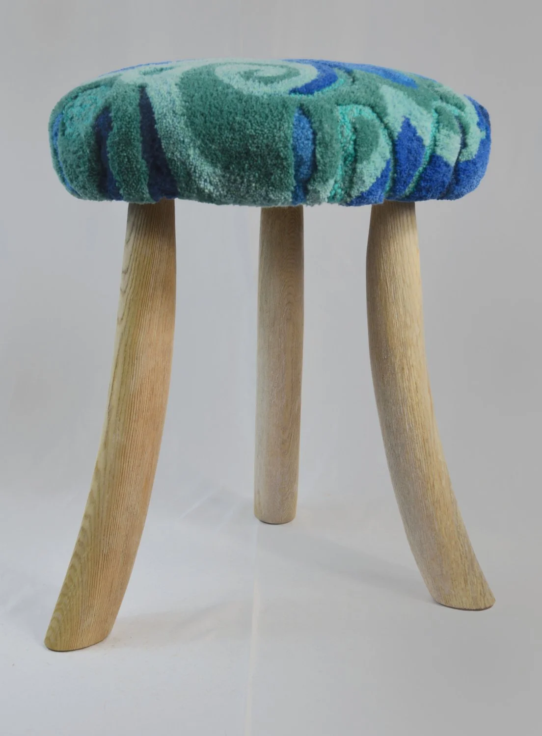 A small round hardwood stool with three wooden legs and a cushioned top covered in a teal and blue patterned fabric.