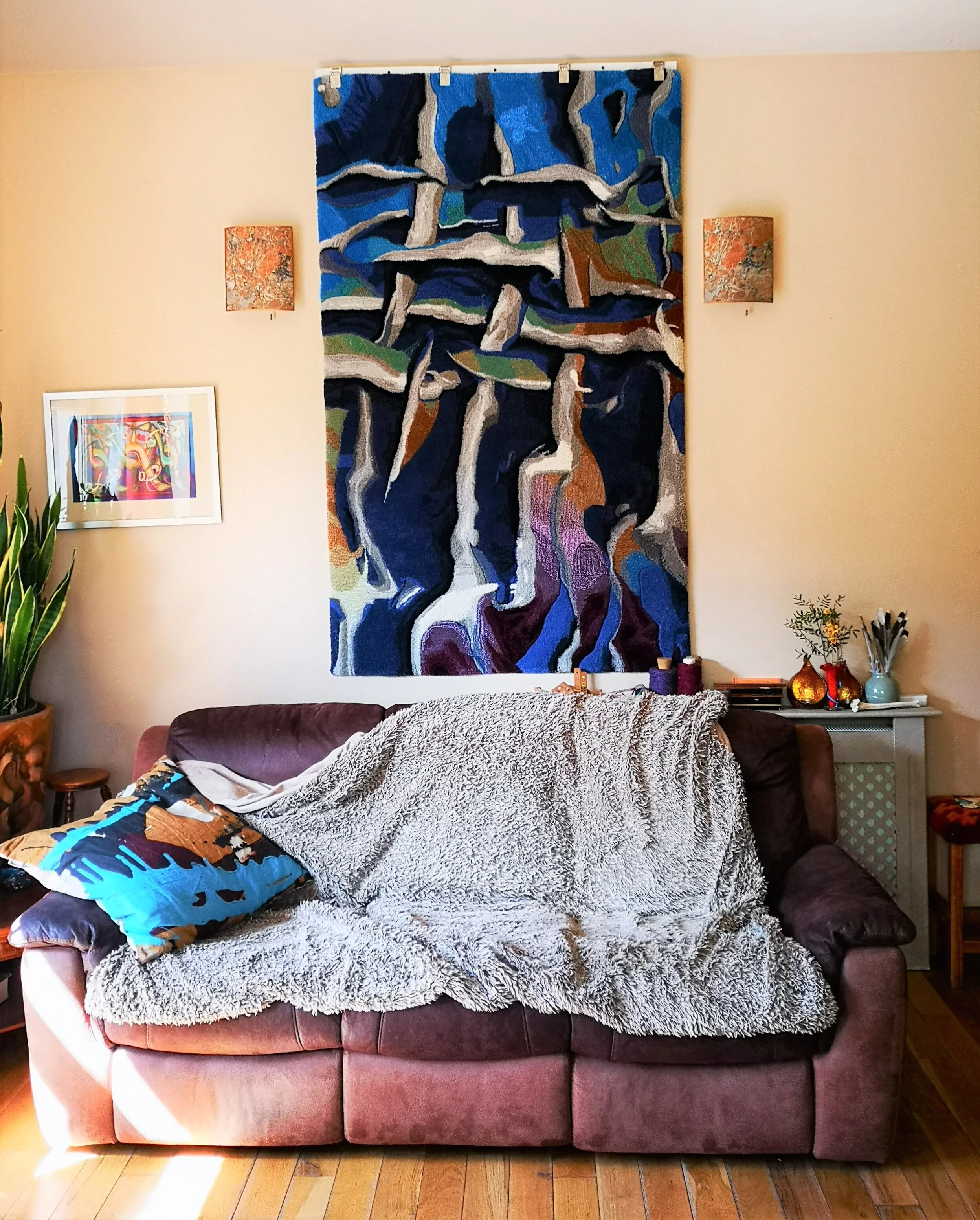 Living room with a brown couch covered in a blanket, a decorative pillow, large colorful abstract wall hanging textile above, small painting on the side wall, potted plant on the left, and a side table with vases..