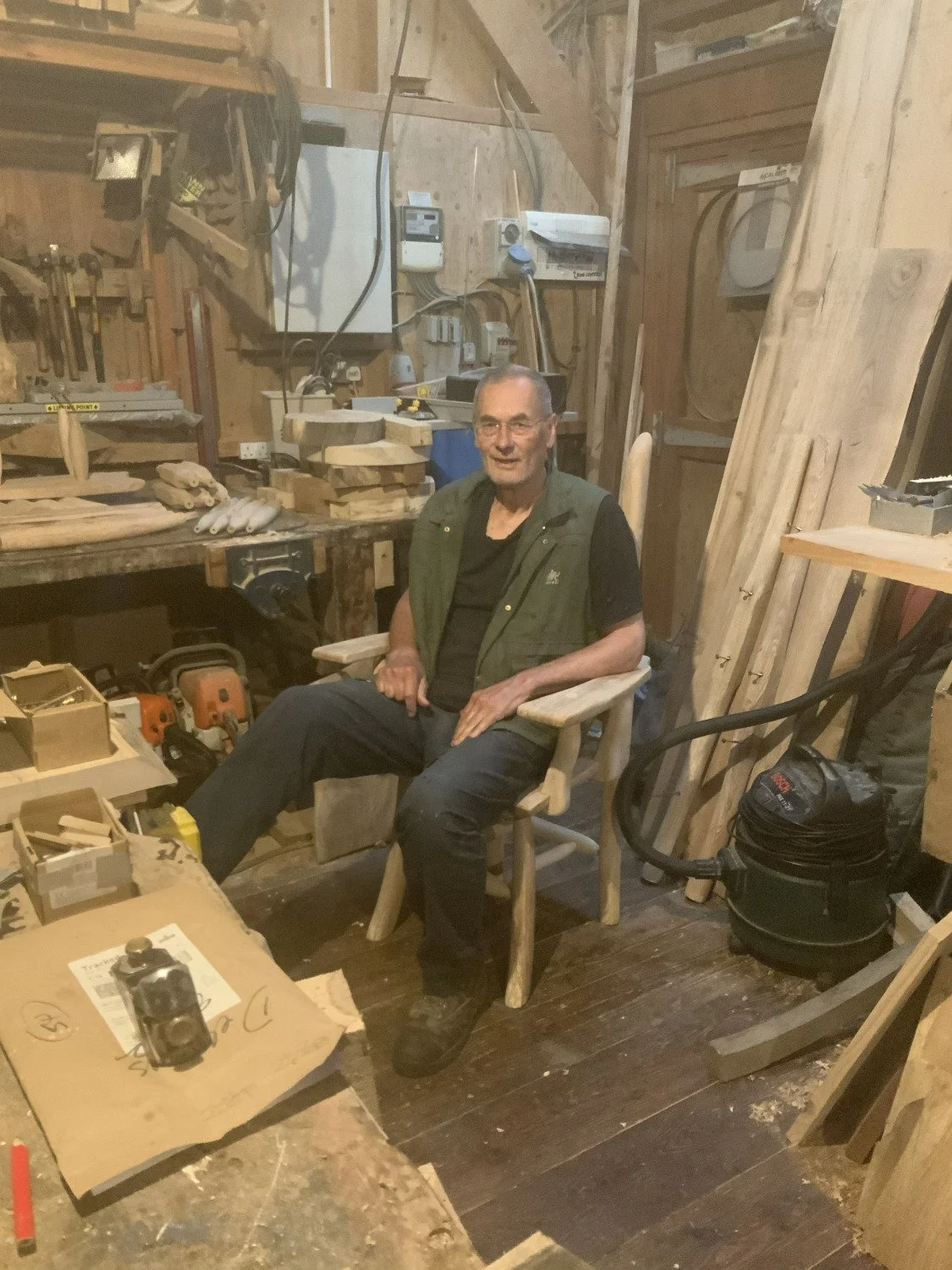 An older man sitting in a wooden workshop, surrounded by wood pieces, tools, and machinery, with a relaxed expression.