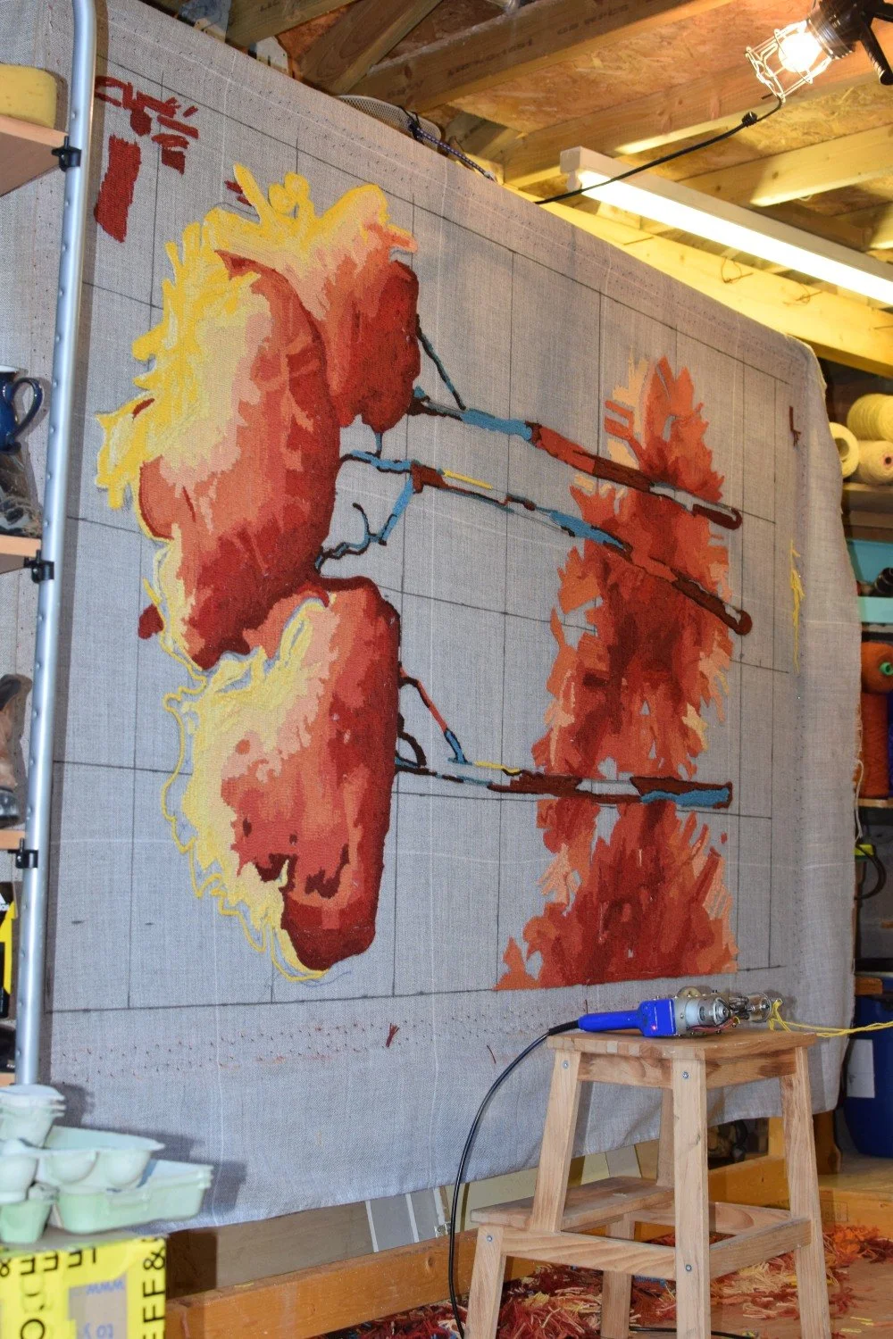 A large hand tufted artwork in progress of a wool pile textile hearth rug, created on a fabric canvas, with work tools and supplies in a workshop setting.