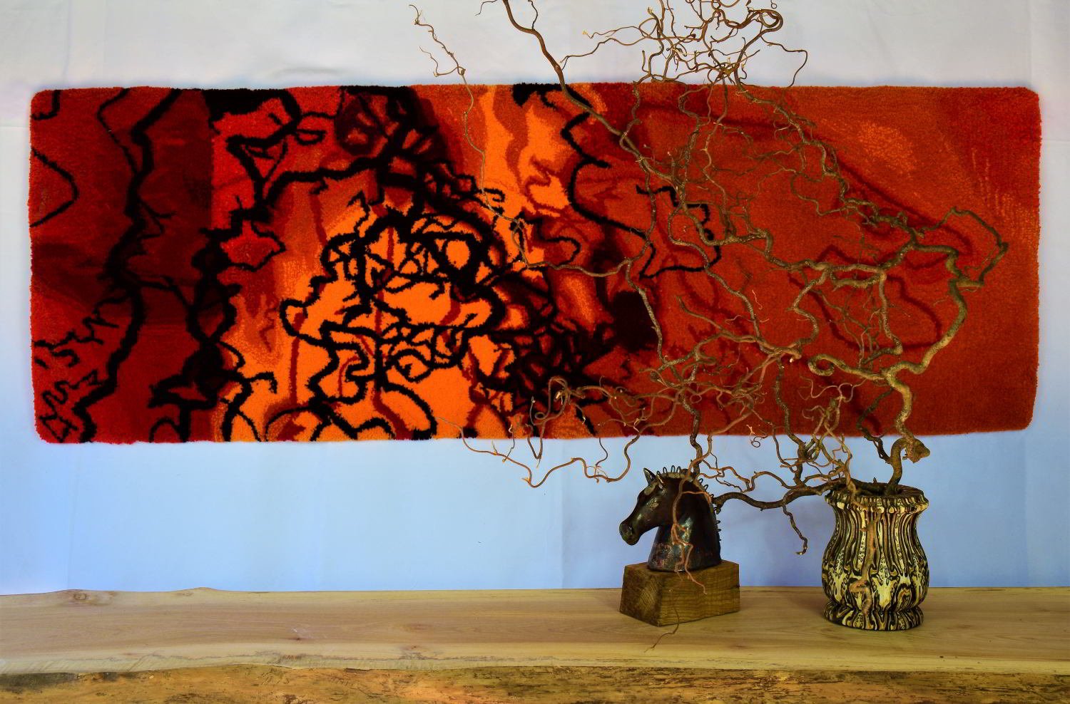Decorative interior display featuring a large abstract red and black textile wall hanging, a twisted wire sculpture resembling a tree or plant, a decorative horse head figurine on a wood base, and a ceramic pot with an ornate black and yellow pattern on a wooden shelf.