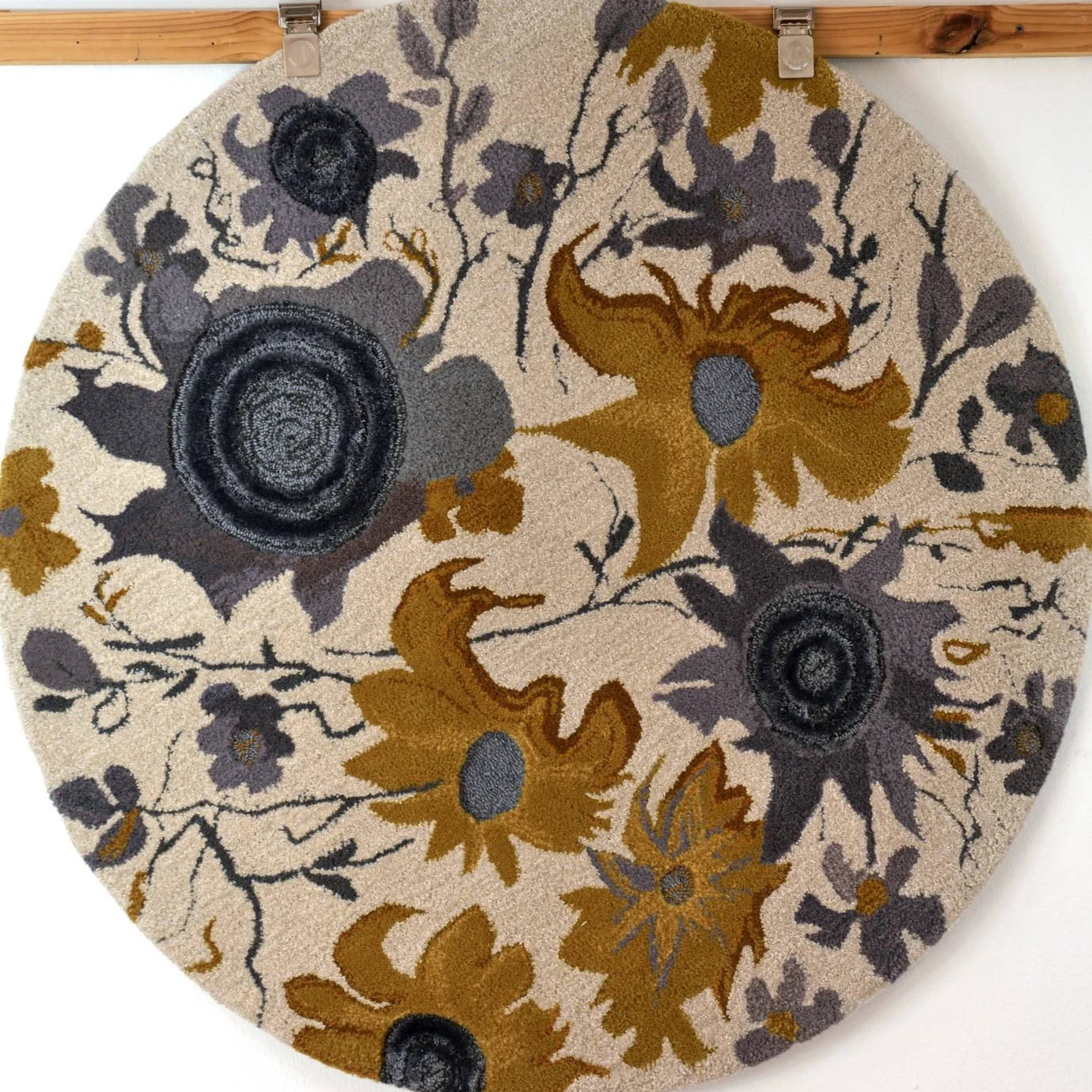 A round floral patterned rug with large flowers in shades of gray, black, mustard yellow, and beige, hanging from two silver clips on a wooden bar.