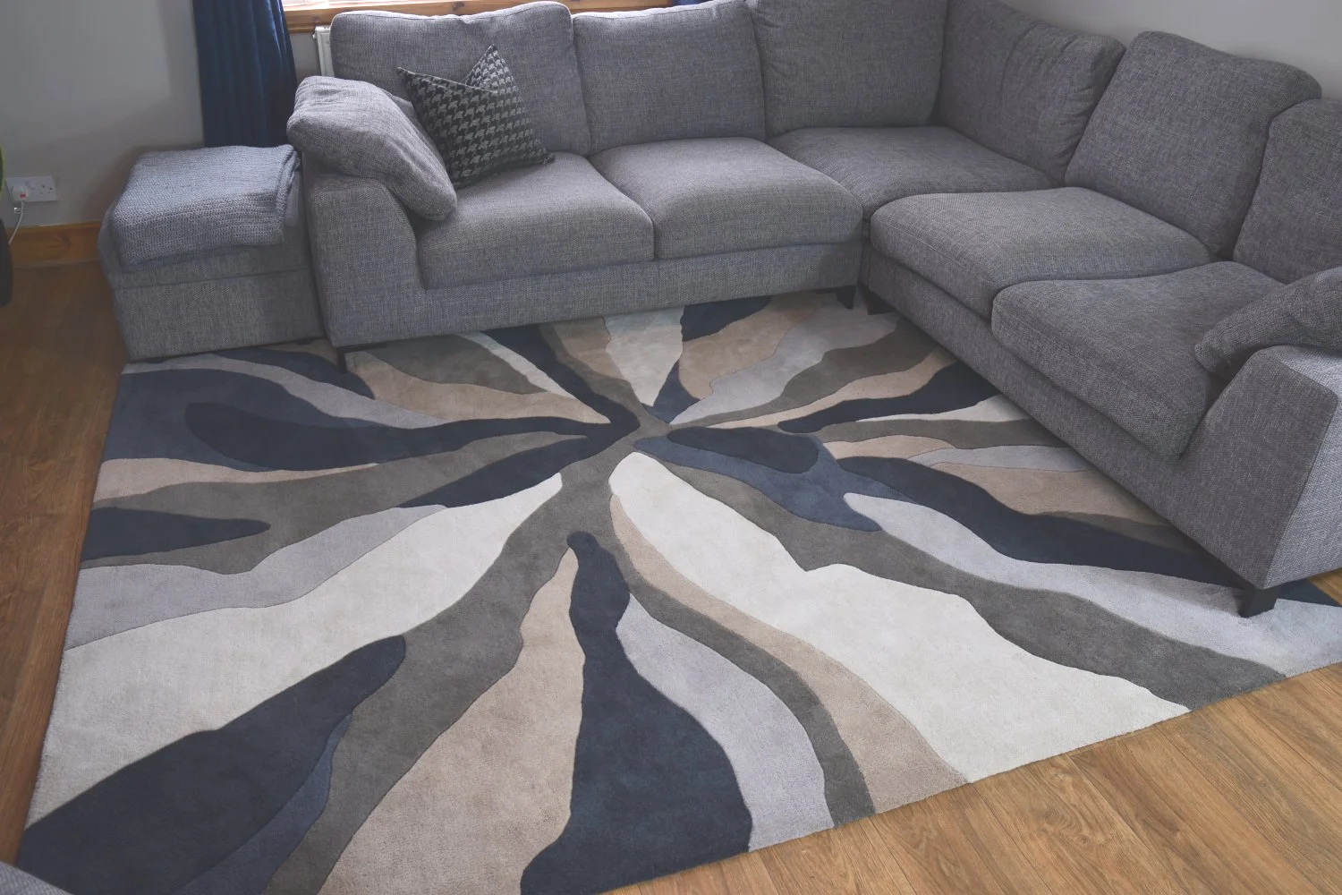A large gray L-shaped sectional sofa with a black-and-white patterned throw pillow, situated on a multicolored abstract area rug with shades of gray, black, beige, and white, on a wooden floor.