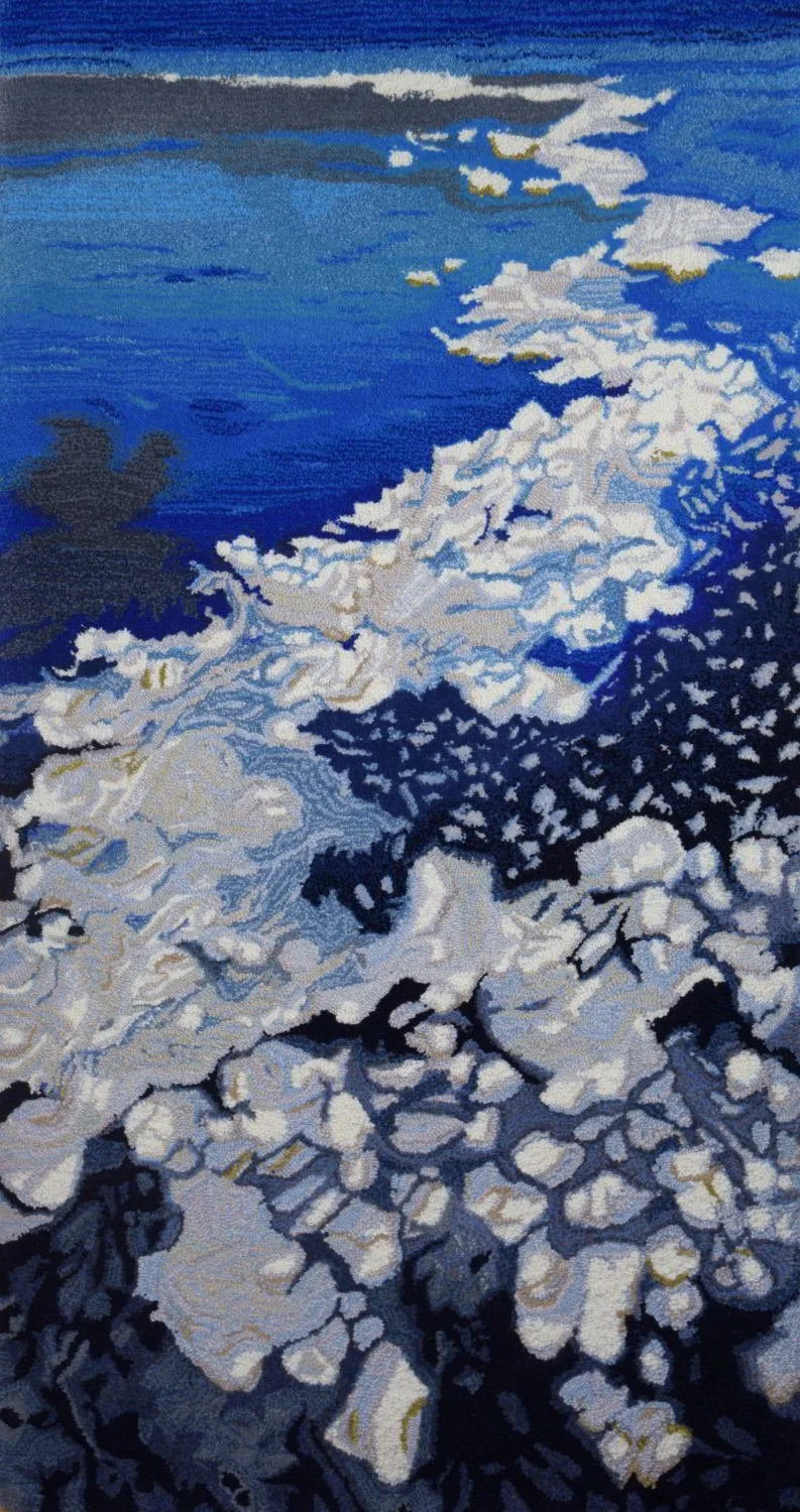 A sea foam hand made wool pile textile of a shoreline with foam and waves crashing onto rocks and sand, under a blue sky.