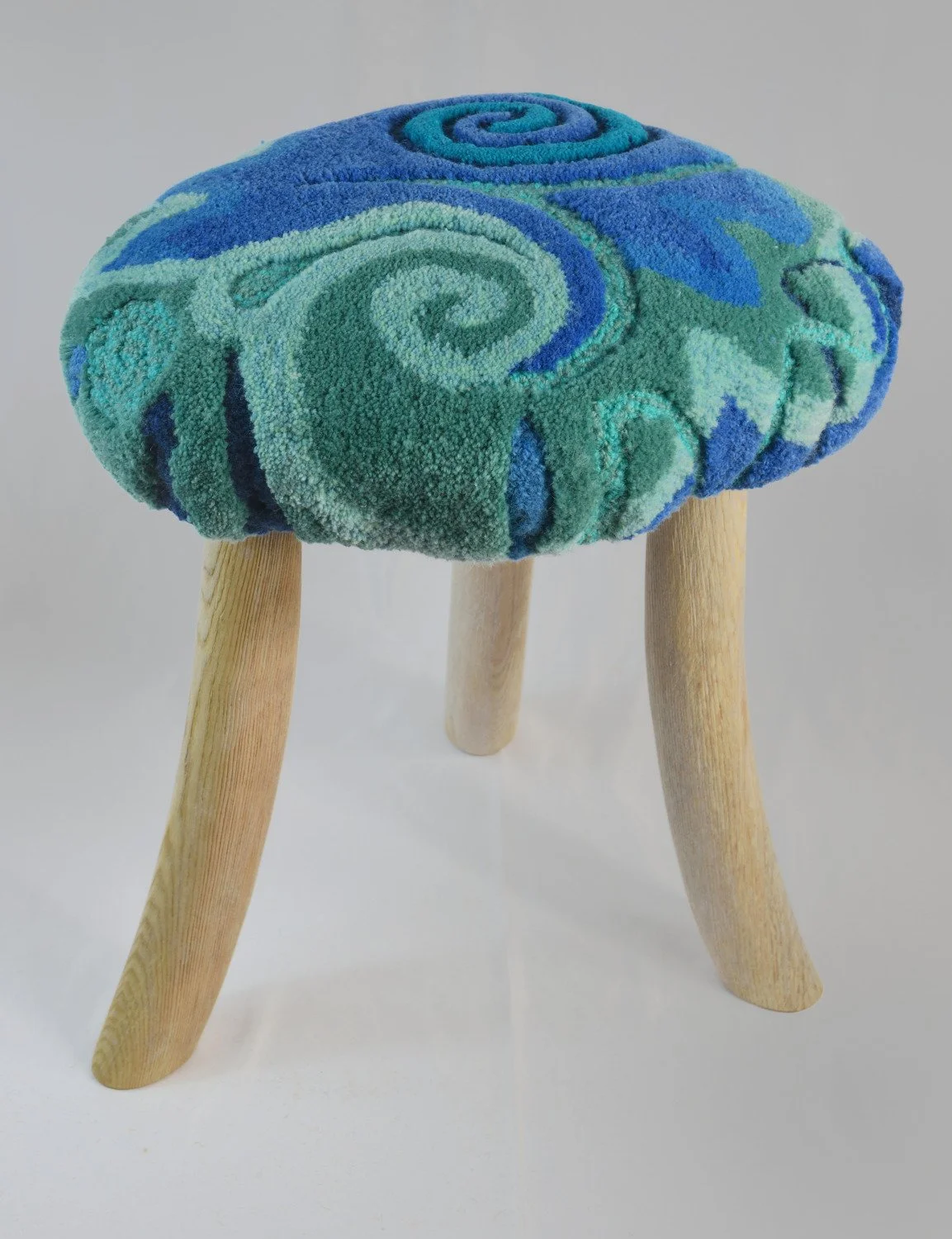 A hard wood stool with a plush, celtic swirl-patterned fabric cushion top in shades of blue and green, with three wooden legs.