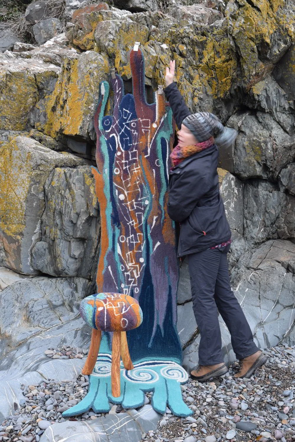 Person in black jacket and pants interacts with outdoor art installation resembling a colorful abstract cactus, with a matching stool, on a rocky beach.