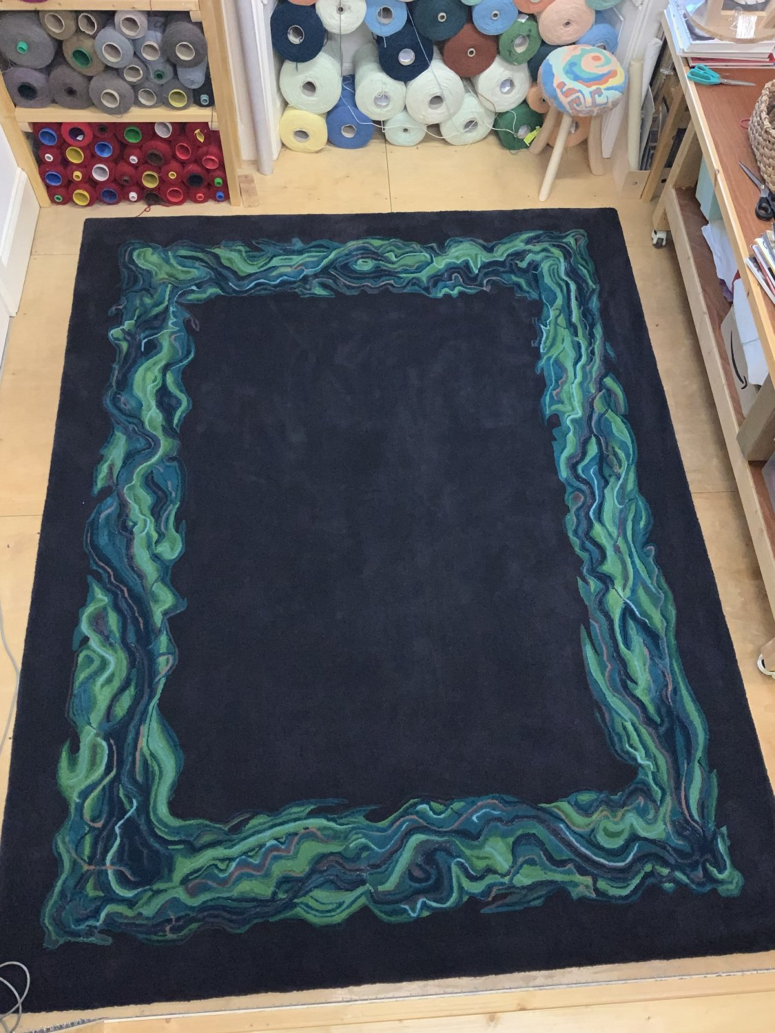 A deep aubergine rug with a colorful abstract border design in shades of green, blue, and purple in the textile artists studio