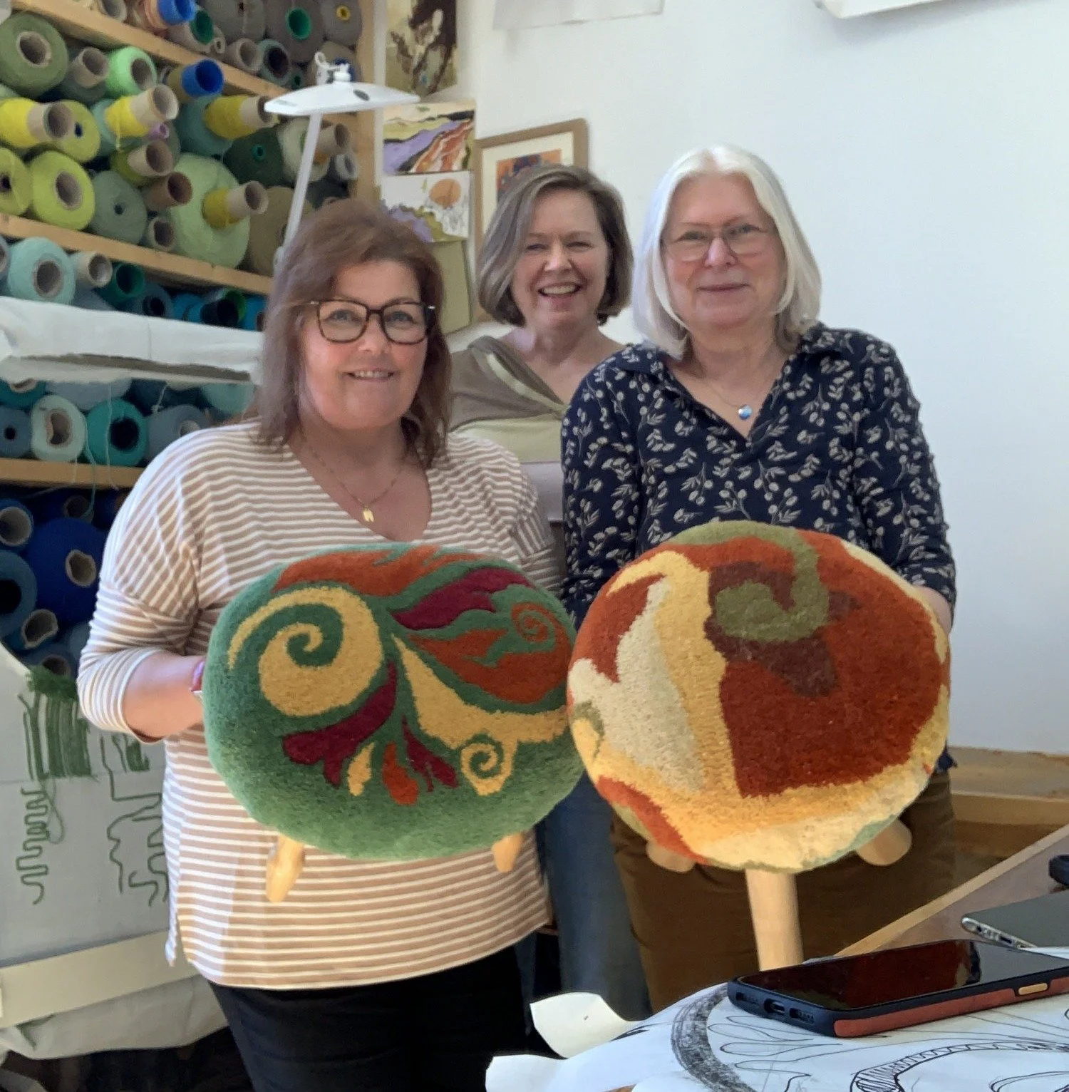 Three women standing indoors, each holding a large, colorful, spherical art piece hardwood stool they have made at a workshop, smiling at the camera. The background includes shelves with rolls of colored yarn or fabric and some framed artwork.