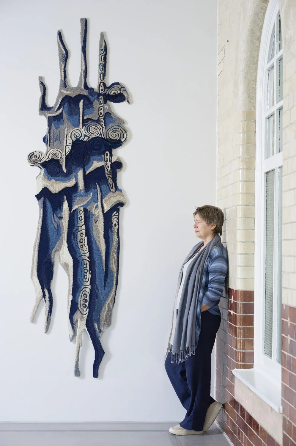 A woman standing and leaning against a brick wall, looking at a large, abstract textile artwork on a white wall.