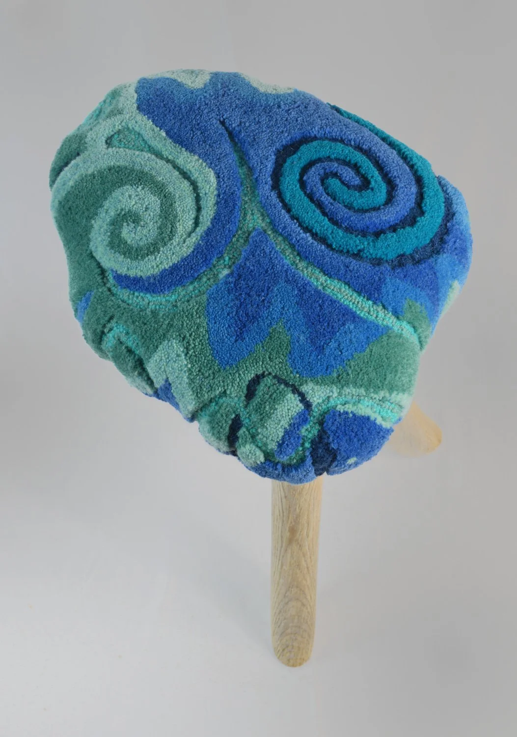A plush hard wood stool with a swirl of blue and green fabric on top, mounted on three hardwood legs.