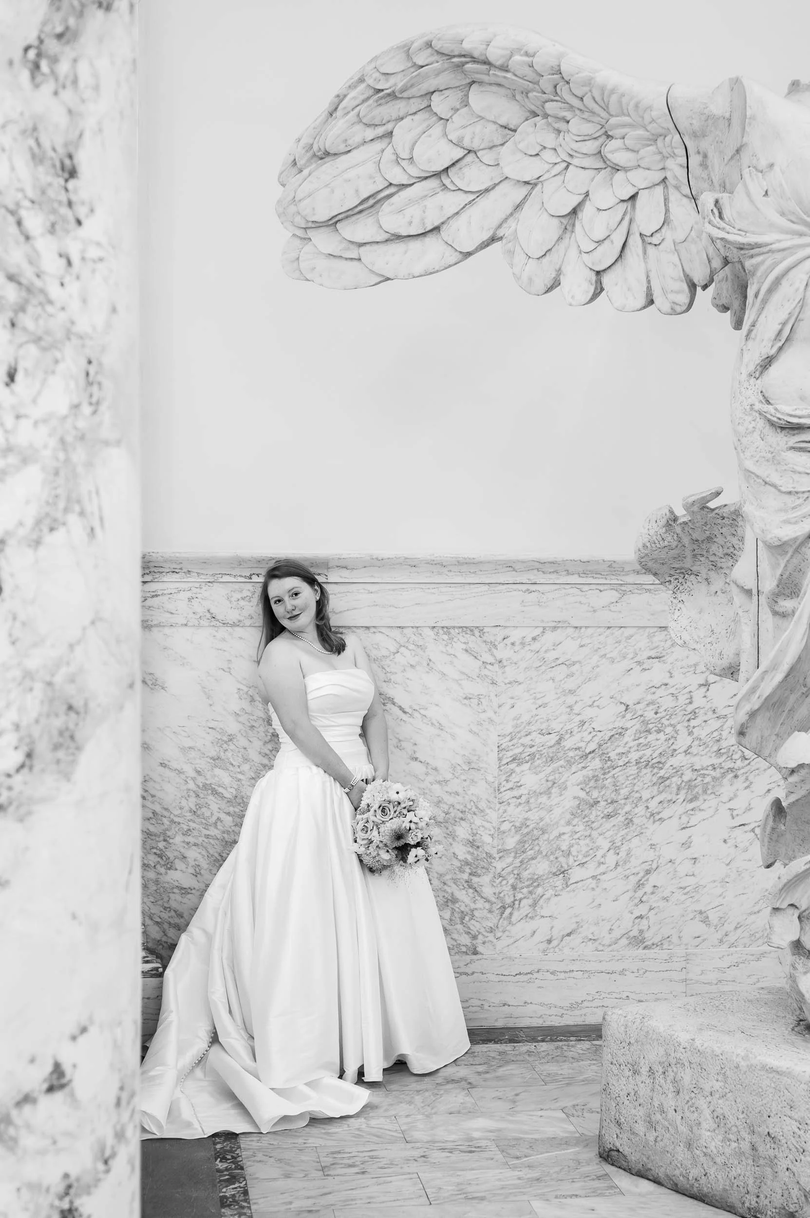 A bride in a strapless wedding gown holding a bouquet, standing in a marble-walled space with classical sculpture and architectural details.