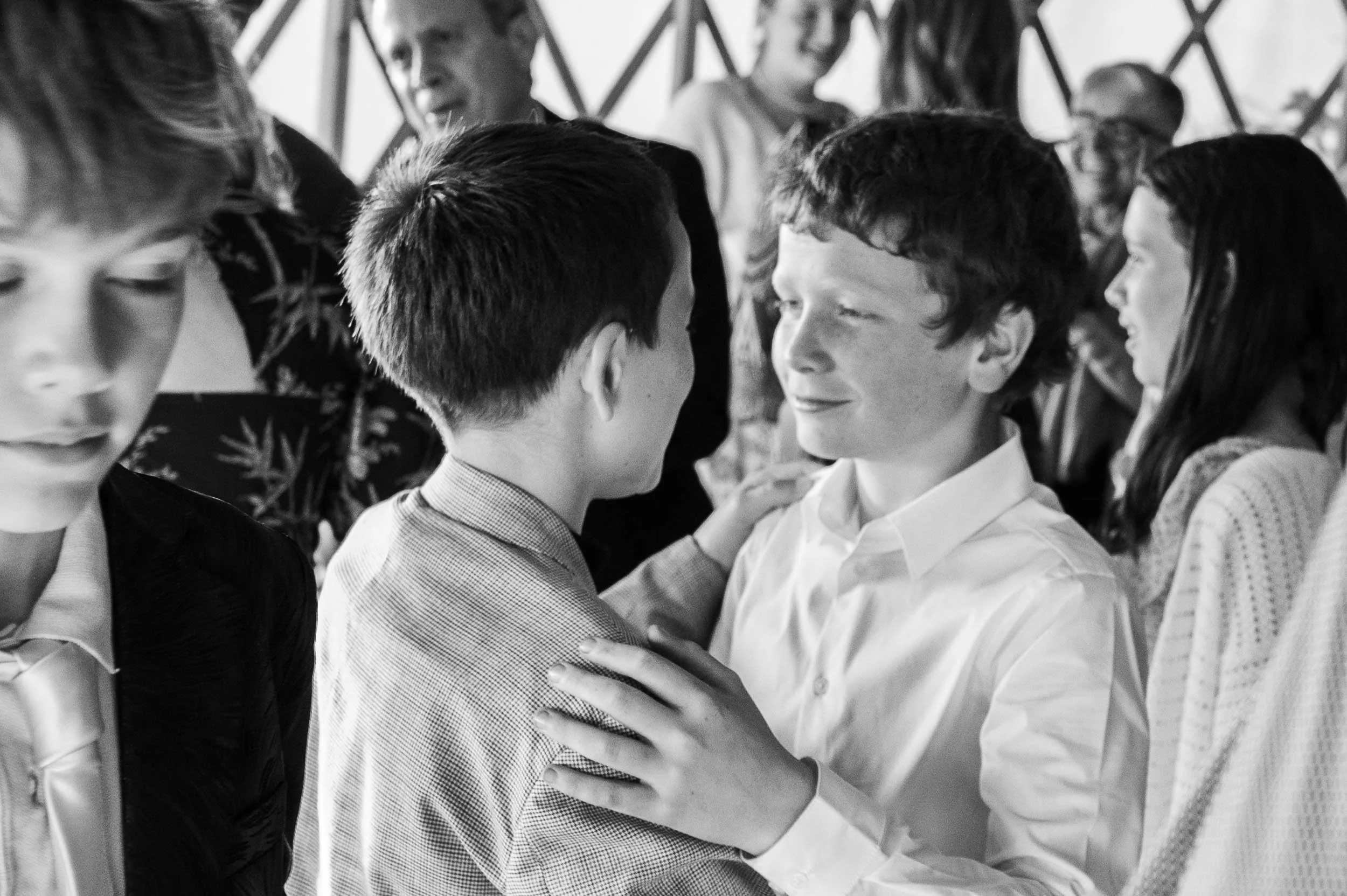 Two boys facing each other and smiling, with a group of people in the background, some talking and some observing, possibly at a social gathering or event.