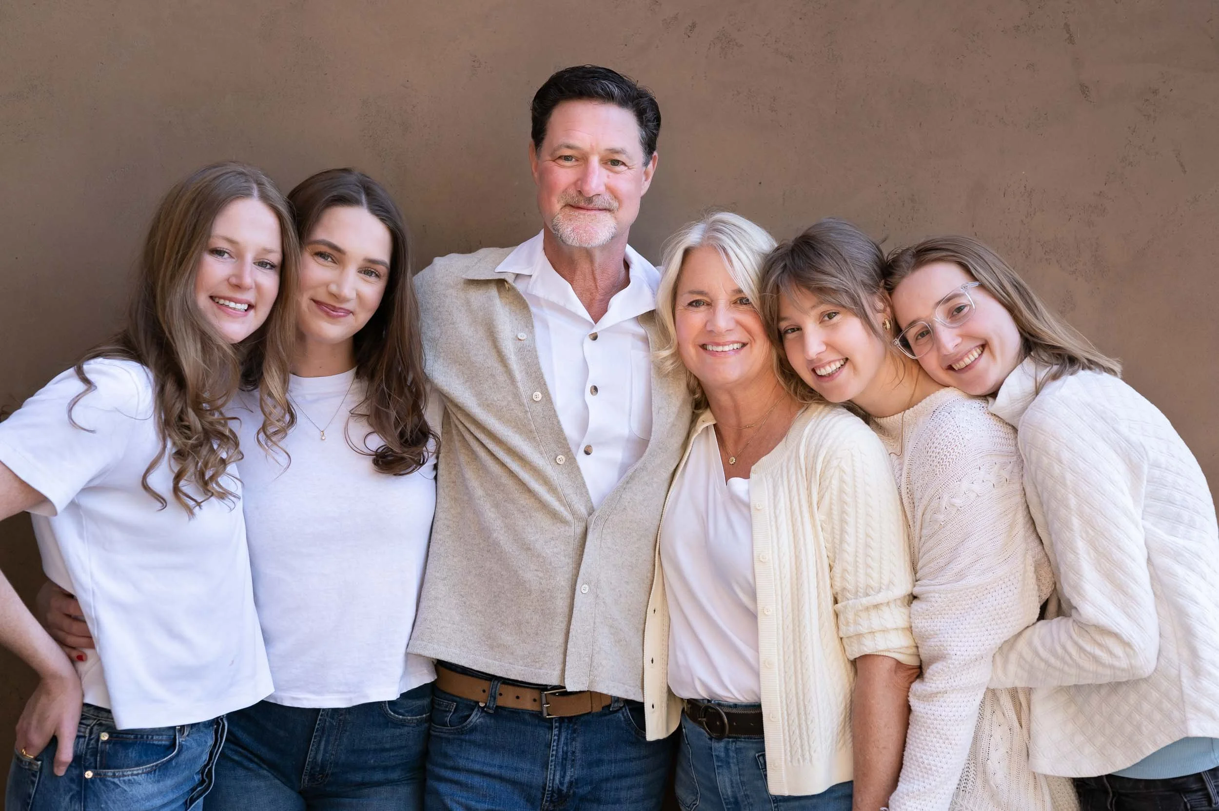 A group of six people, including three young women, a middle-aged woman, a middle-aged man, and a young woman, standing together outdoors against a brown wall, smiling and posing for the camera.