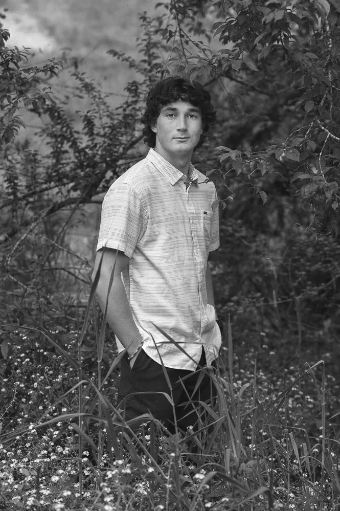 A young man standing outdoors among tall grass and plants, surrounded by leafy trees in a natural setting.