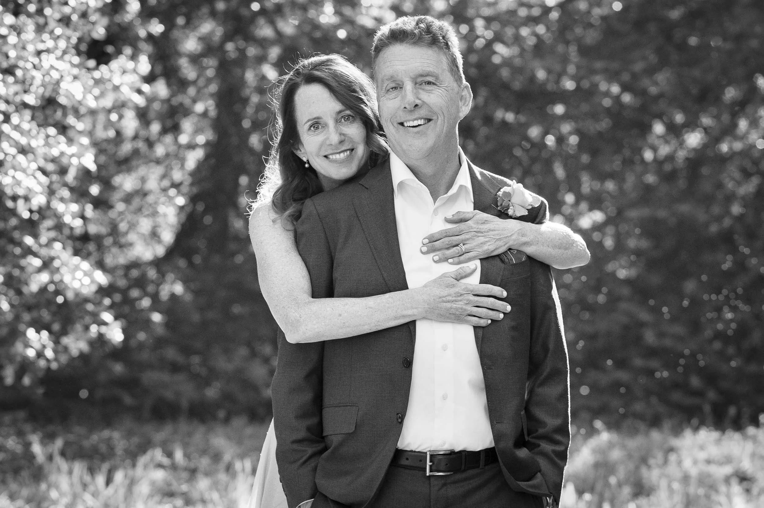 A black and white photo of a happy couple outdoors, with the woman hugging the man from behind, both smiling. The man is wearing a suit jacket and a white shirt, while the woman is in a sleeveless dress, with her arms around his shoulders.