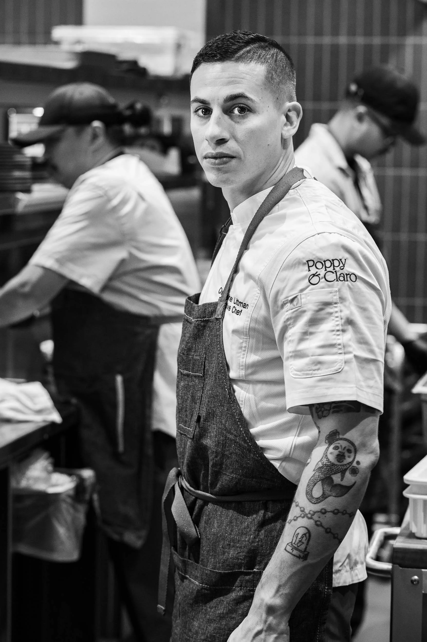 A young male chef with tattoos on his arm, wearing a white chef's coat and a dark apron, looking at the camera in a busy kitchen with other chefs working in the background.