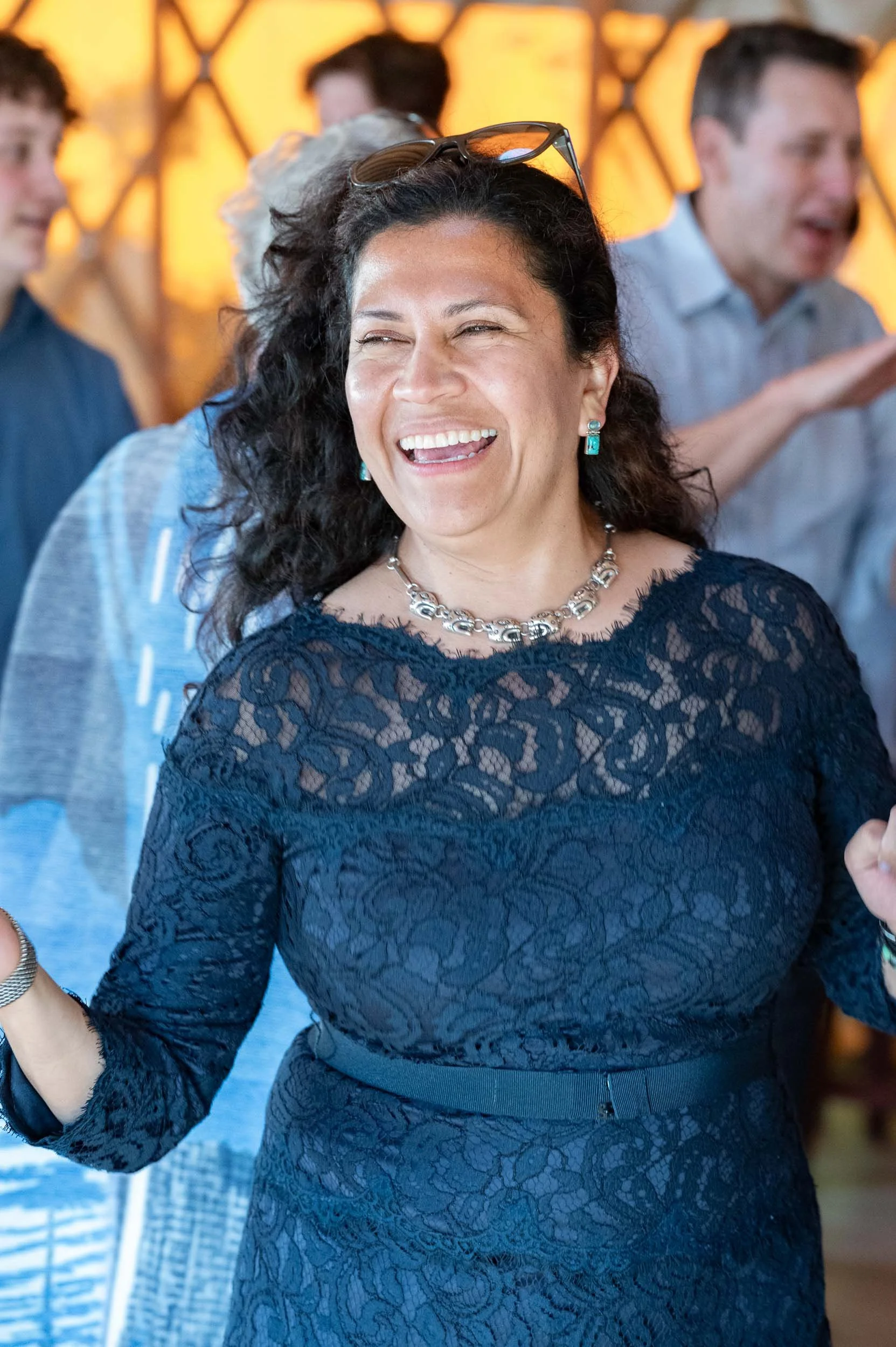 A woman with dark, curly hair laughing at a social gathering, wearing a navy lace dress, jewelry, and sunglasses on her head, with blurred people in the background.