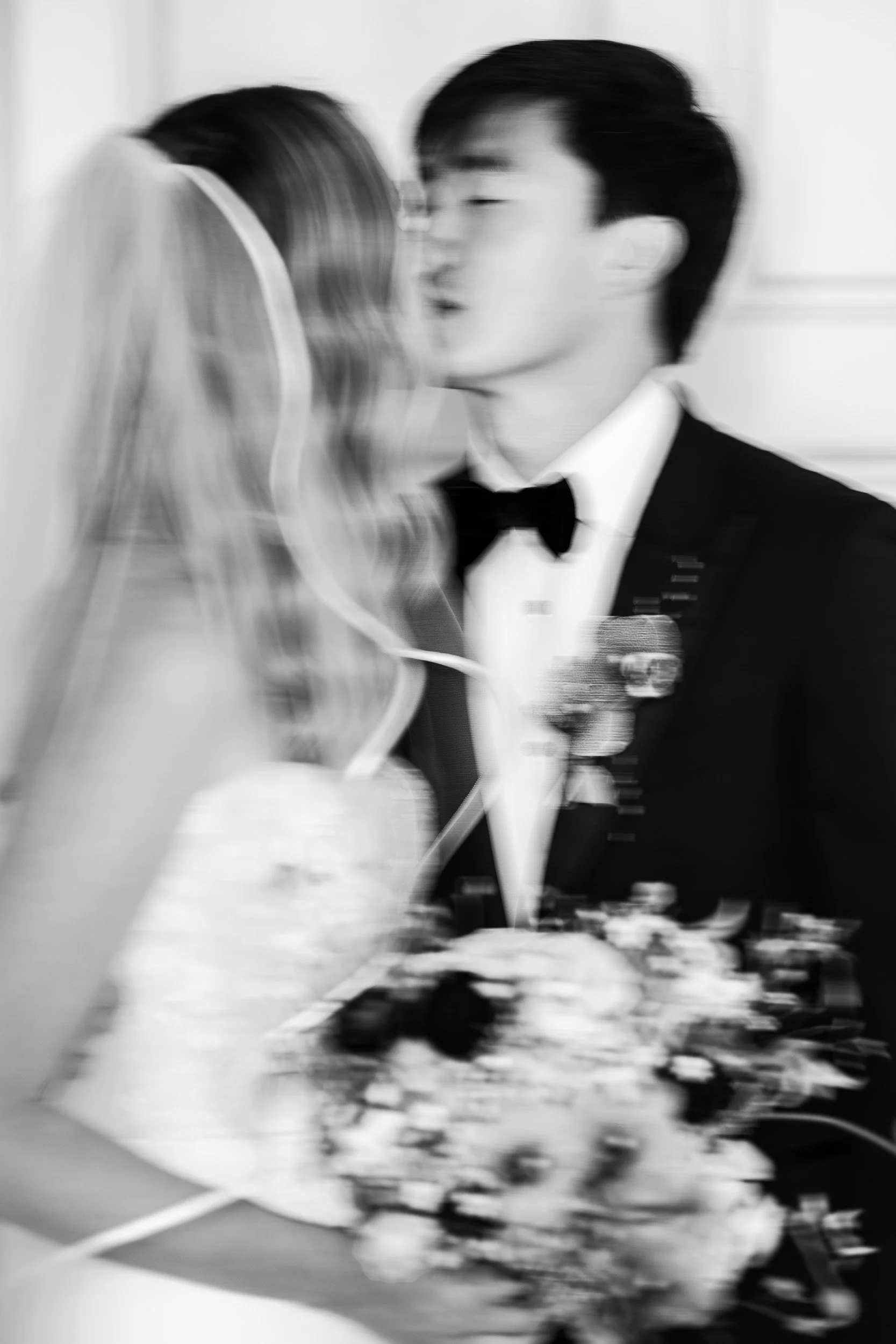 A black and white photo of a man and woman, seemingly about to kiss, blurred due to motion. The man is wearing a tuxedo with a bow tie, and the woman appears to be in a wedding dress with a veil.